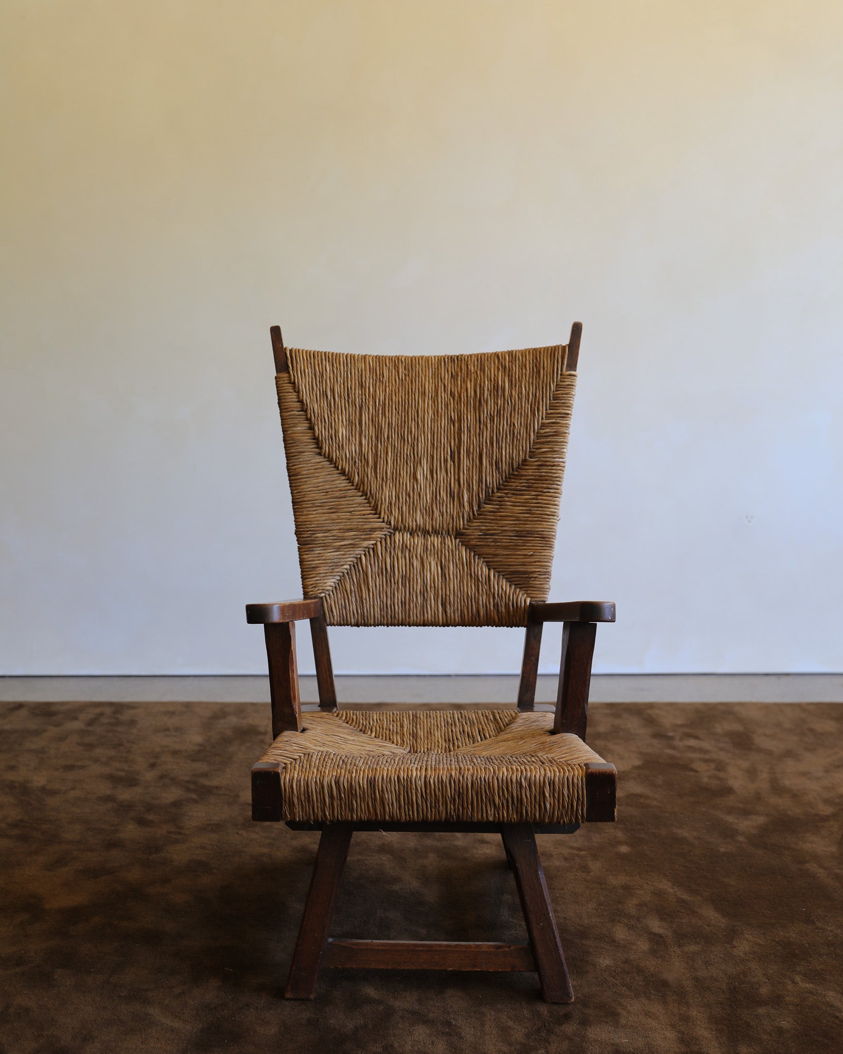 A mid-century modern wooden armchair with a woven natural-fiber seat and back, set against a plain beige wall and textured brown floor.