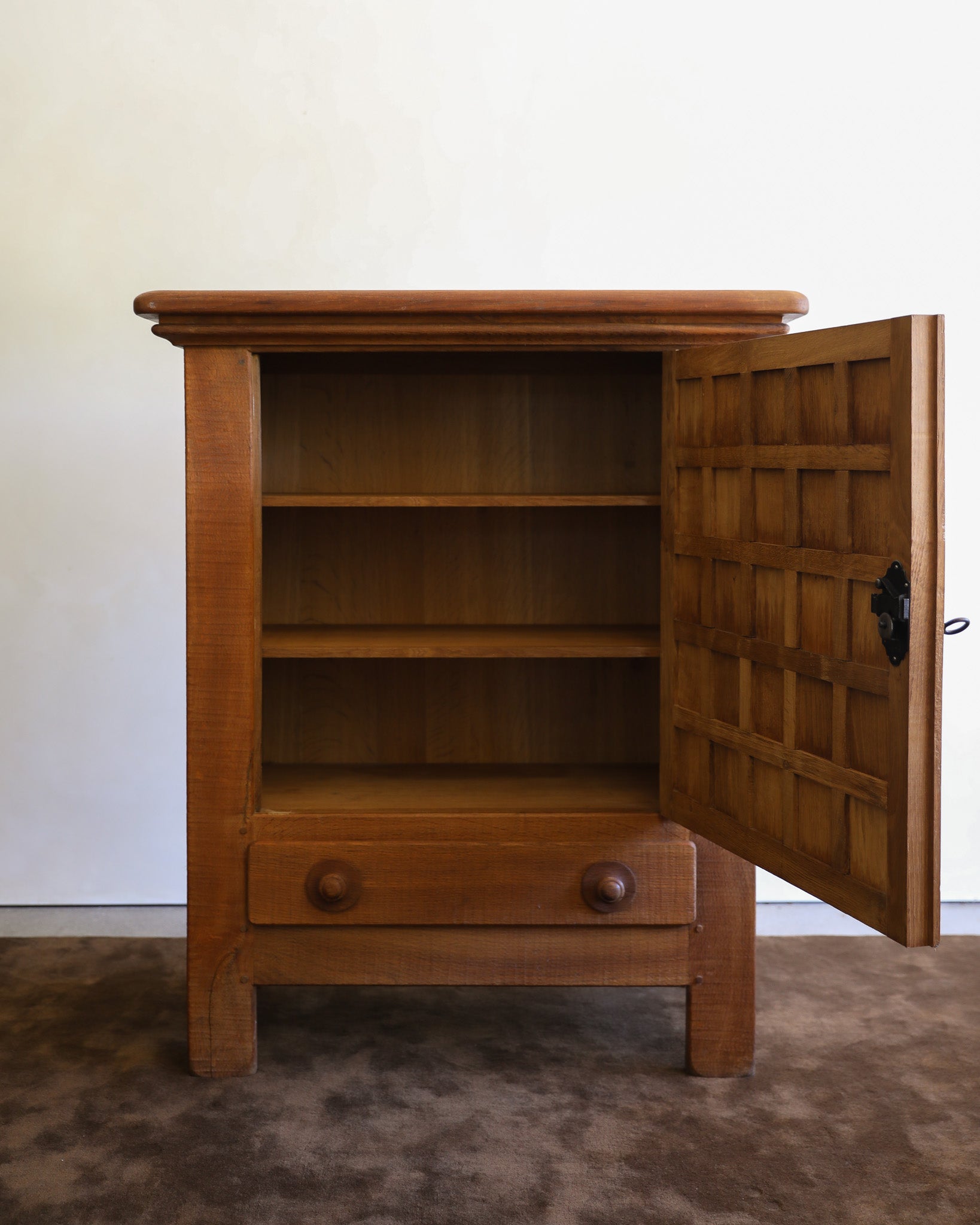 A wooden cabinet with an open door, showing three interior shelves and a single drawer with round knobs, set against a plain off-white wall and brown carpet.