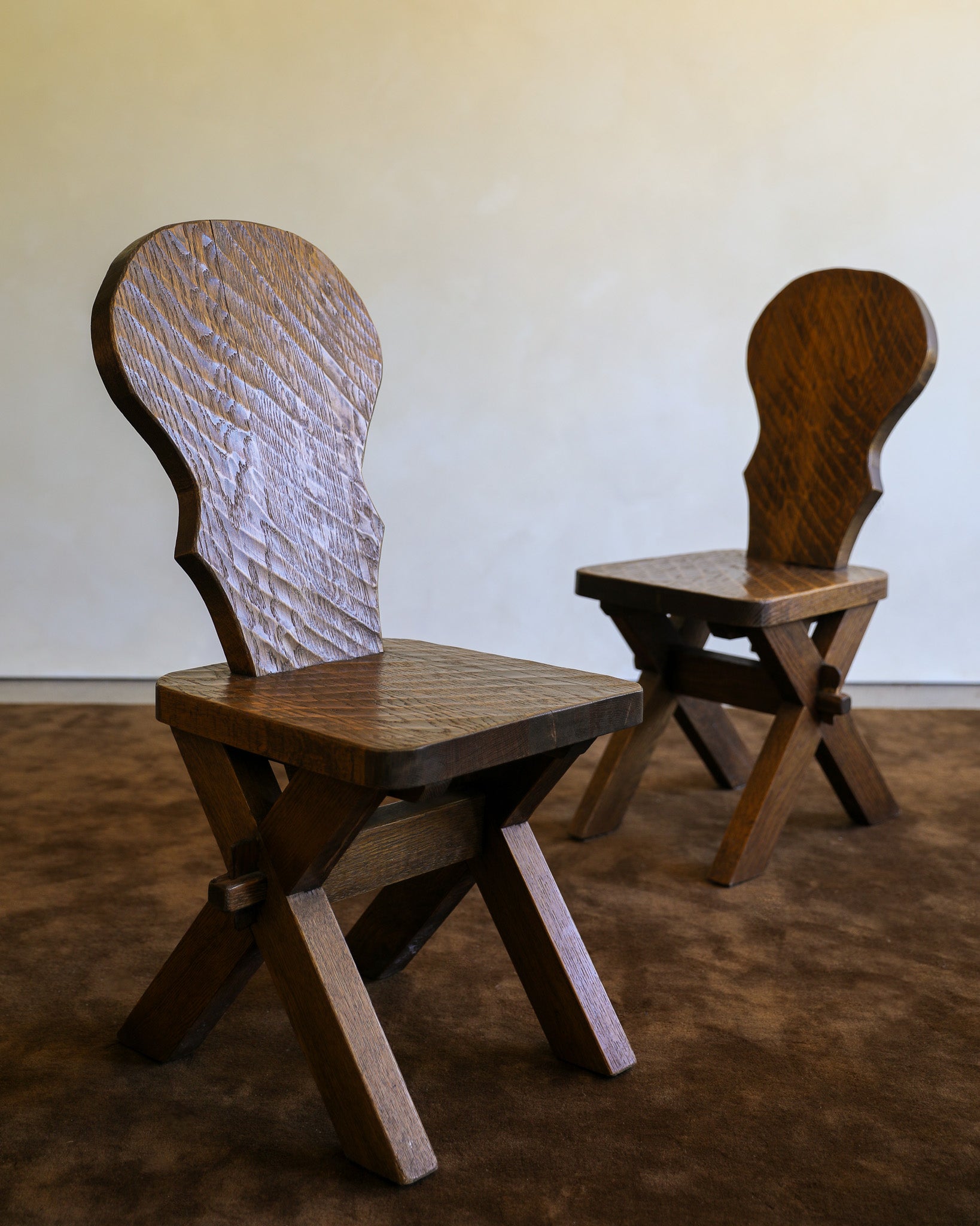 Two rustic wooden chairs with carved backs, shown on a brown carpet against a plain beige wall.