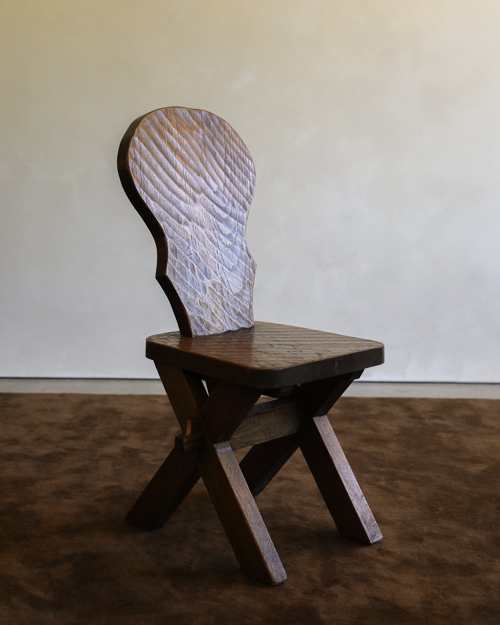 A rustic wooden chair with a square seat, crossed X-shaped legs, and a uniquely carved, textured backrest, set against a plain beige wall and brown floor.