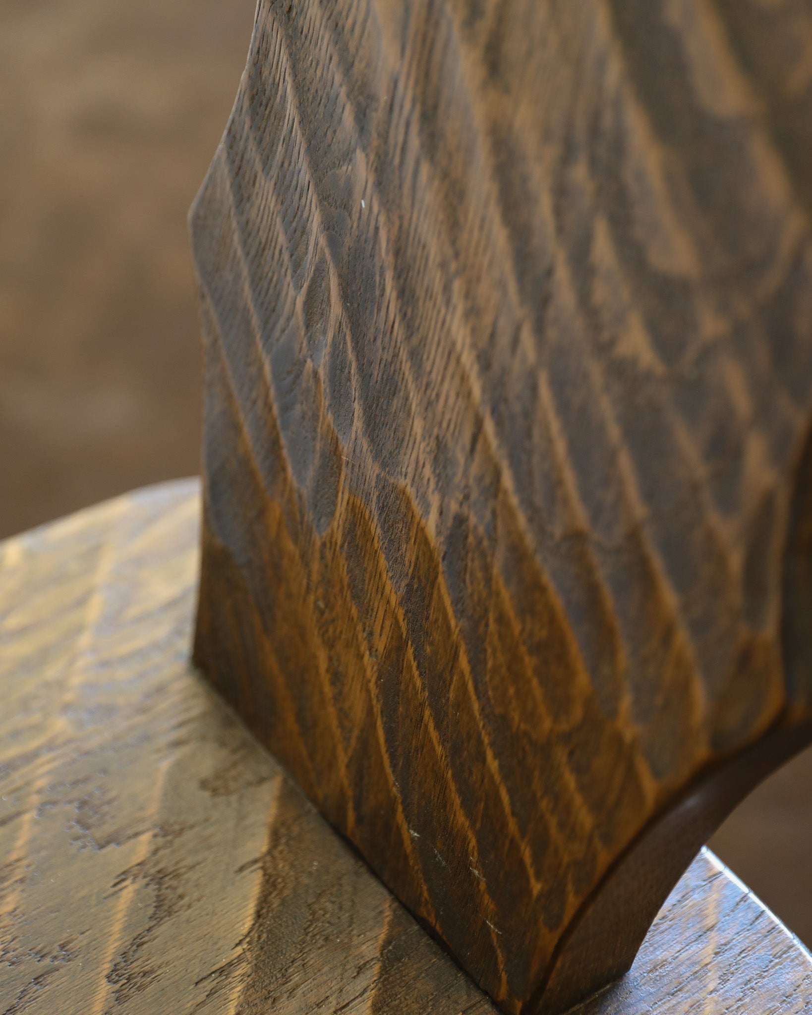 Close-up of a dark brown wooden chair with prominent grain texture, resting on a matching wooden surface with a blurred brown background.
