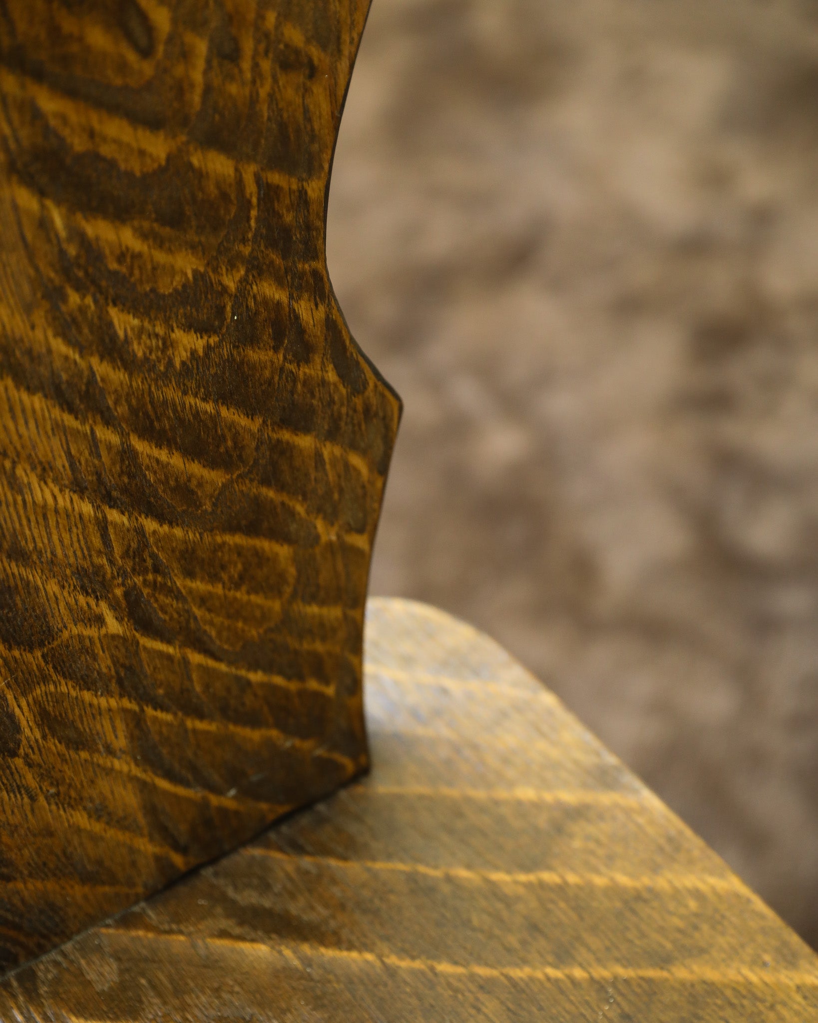 Close-up detail of a rustic wooden chair showing the corner joint between the backrest and seat, with a rich dark brown wood grain finish and a blurred neutral background.