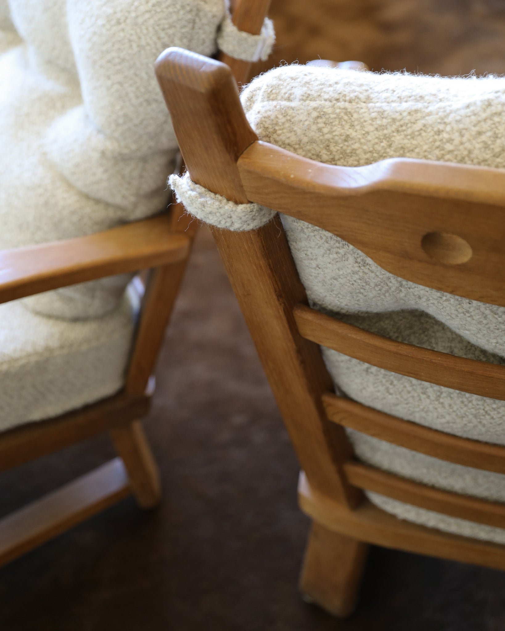 Close-up of a wooden armchair with cream bouclé cushions, showing the curved wooden backrest and seat detail against a dark brown floor.