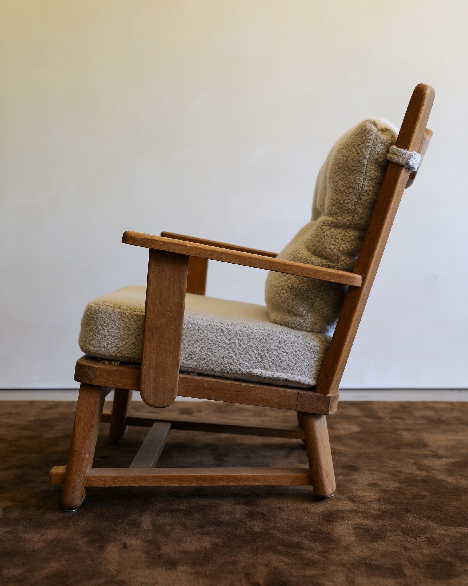 A mid-century modern wooden chair with a textured beige bouclé seat cushion and a matching back cushion, set against a plain off-white wall and dark brown carpet.
