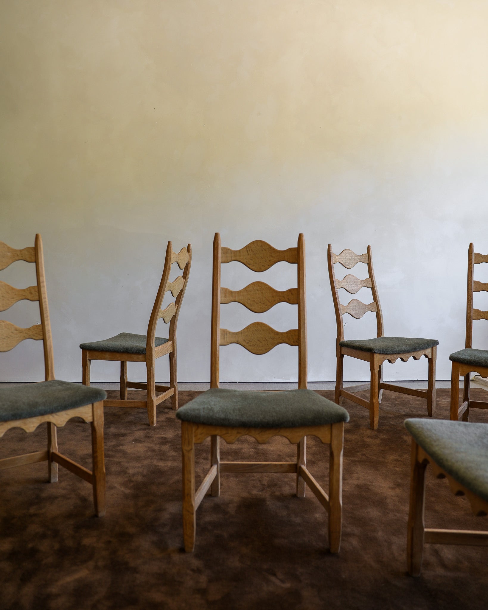 A set of wooden razor-back dining chairs with upholstered seats in muted green, arranged on a dark brown textured rug against a plain beige wall.