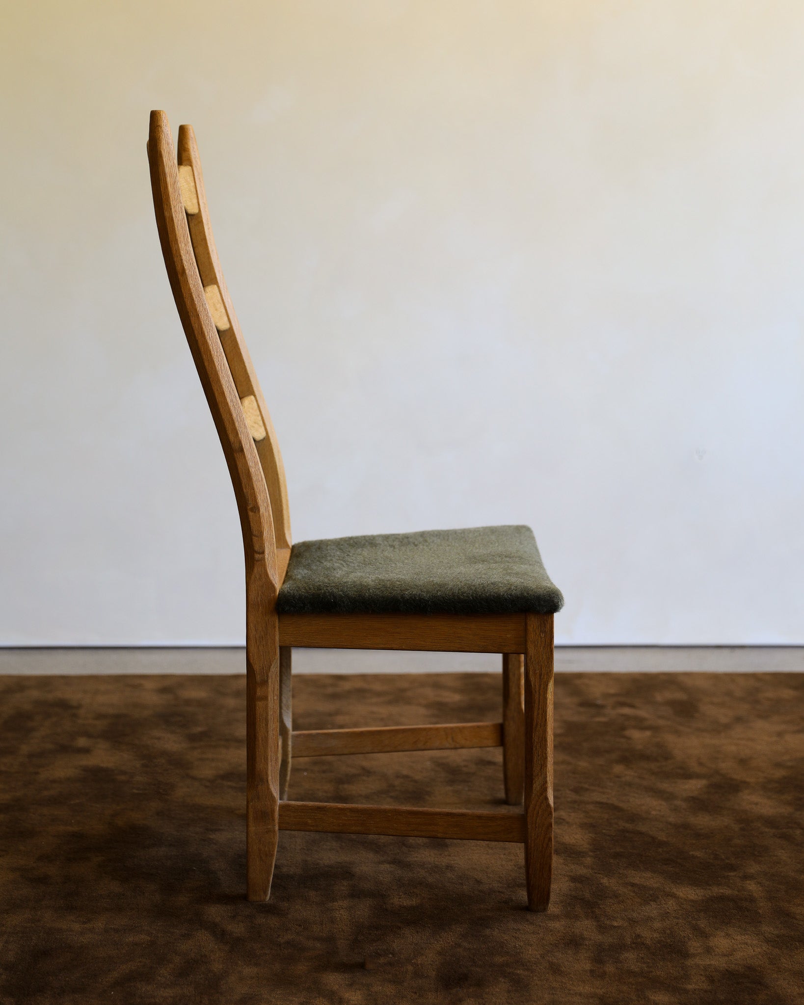 A wooden dining chair with a light oak ladder back and a muted green upholstered seat, shown in a minimalist room setting with a brown rug and a plain off-white wall.