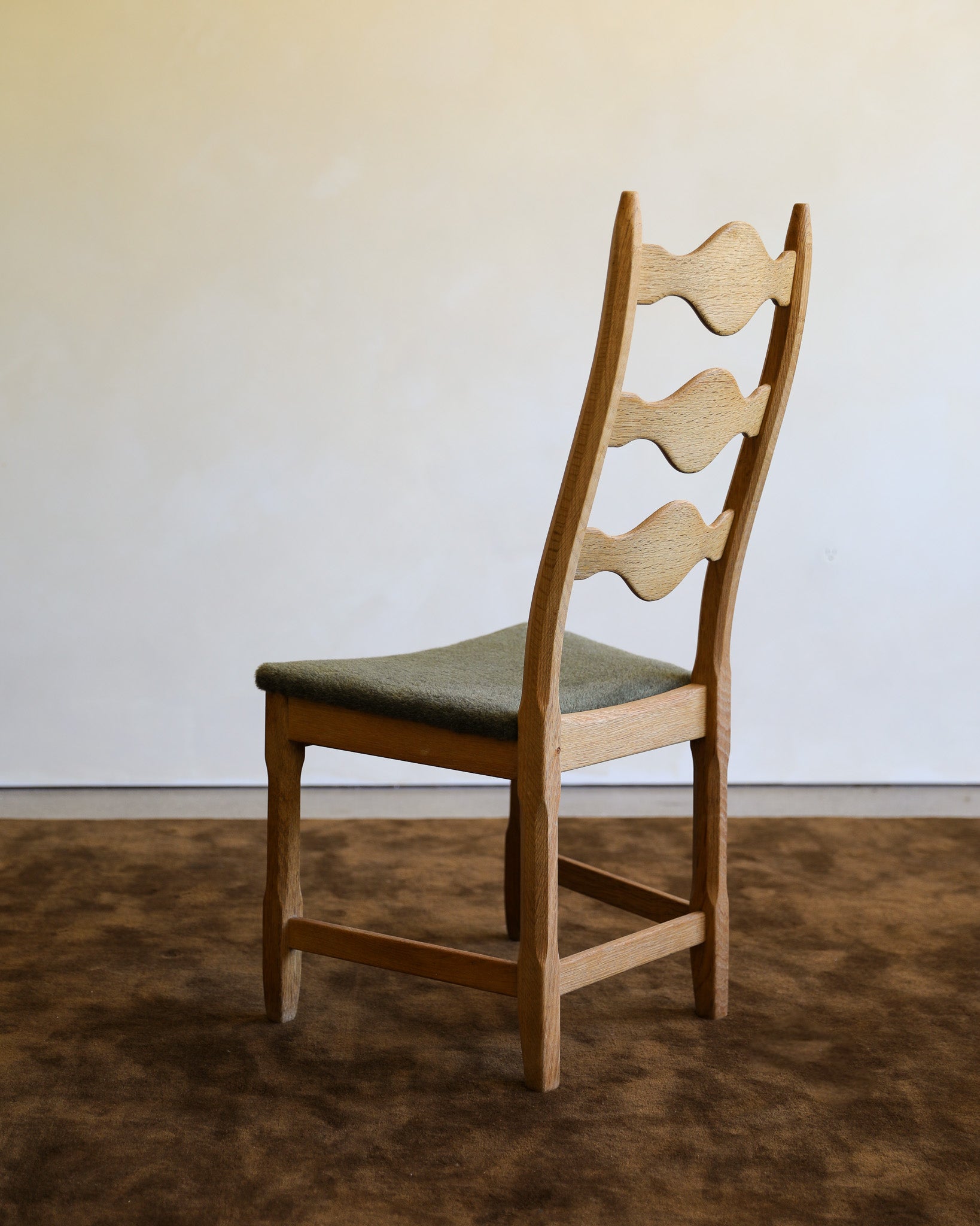 A wooden dining chair with a light oak ladder back and a muted green upholstered seat, shown in a minimalist room setting with a brown rug and a plain off-white wall.