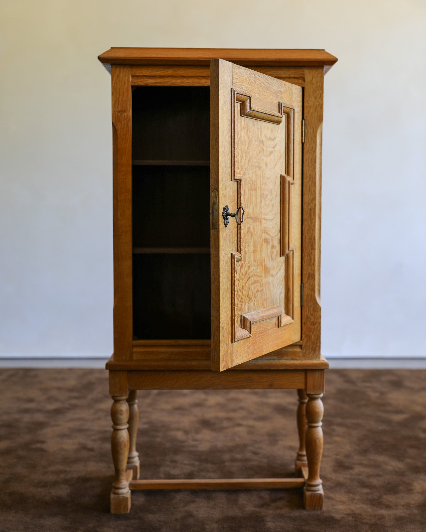 A small wooden cabinet with one door open, showing shelves inside. The cabinet has a warm medium-brown wood finish and stands on turned legs against a plain light wall and brown floor.