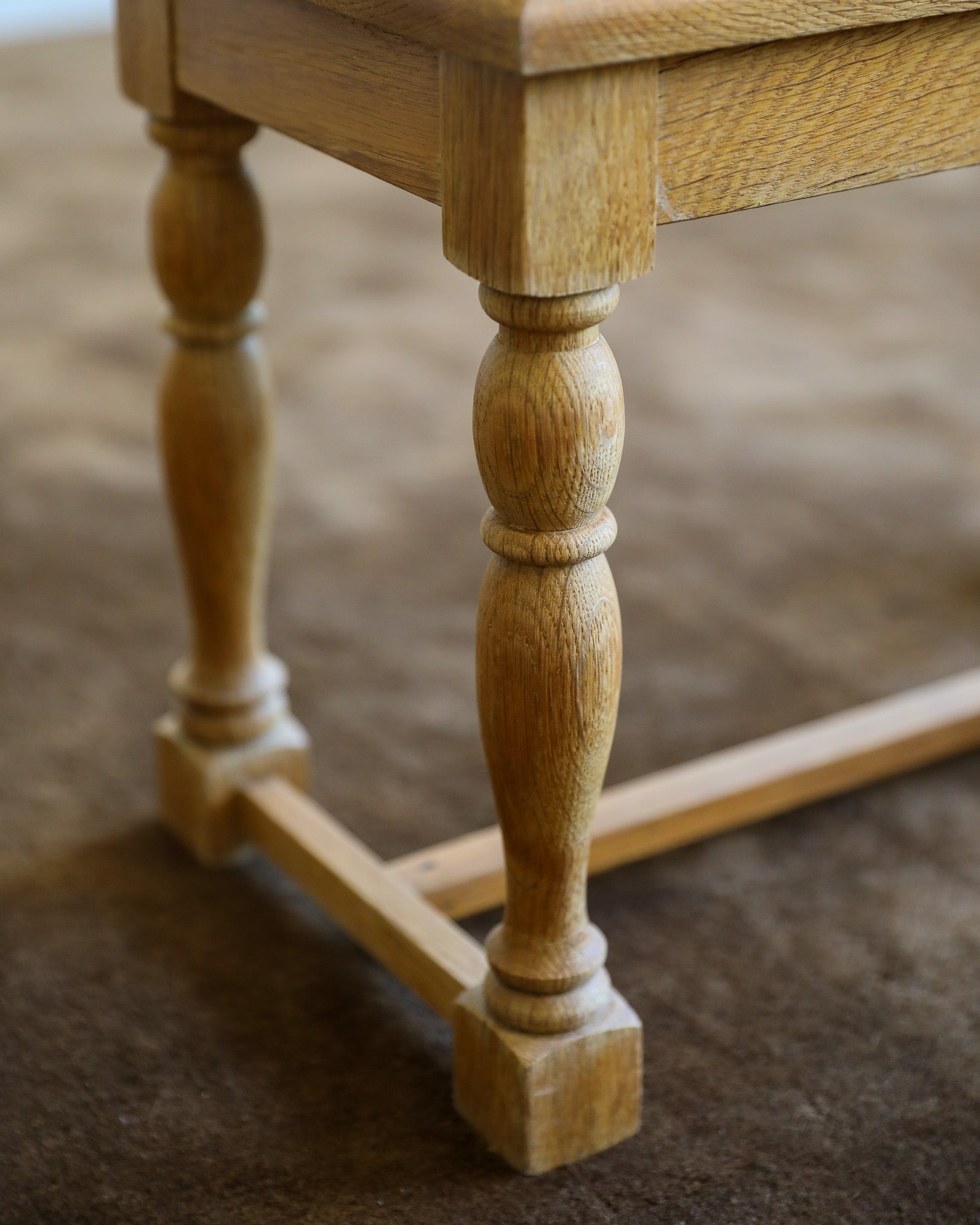 Close-up of a light natural oak wooden stool or bench leg with a turned spindle design and a stretcher bar, resting on a brown carpet.