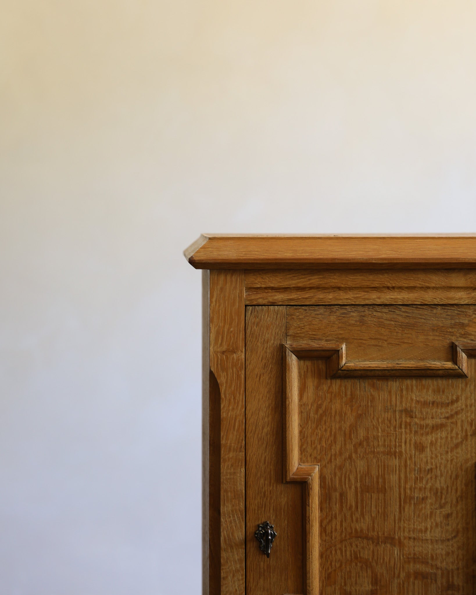 Close-up of a wooden cabinet corner with a warm medium-brown oak finish and a small dark metal keyhole, set against a plain off-white wall.