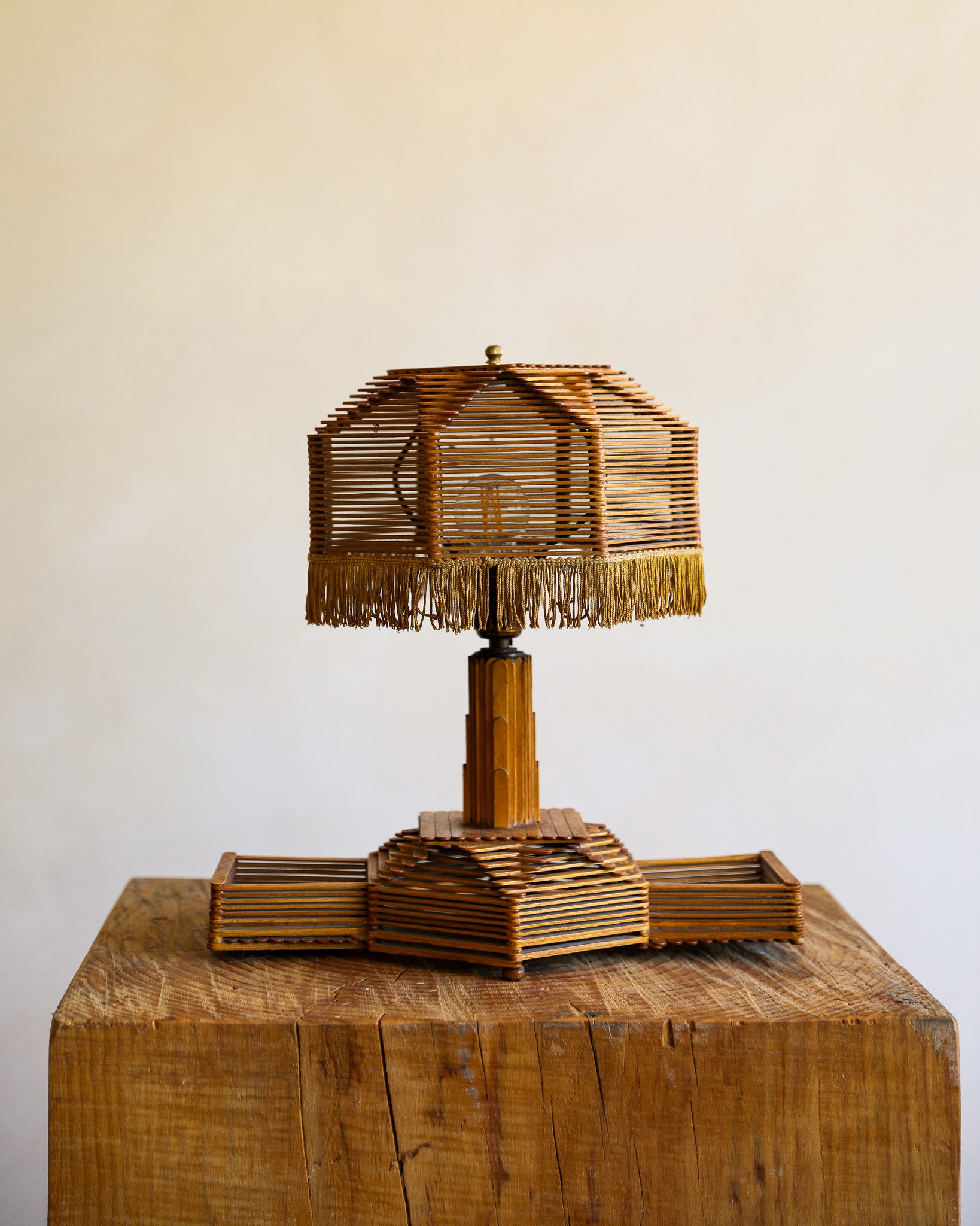 A rustic table lamp crafted from layered popsicle sticks, featuring a fringed shade and a geometric pedestal base, displayed on a large wooden block against a plain light beige wall.