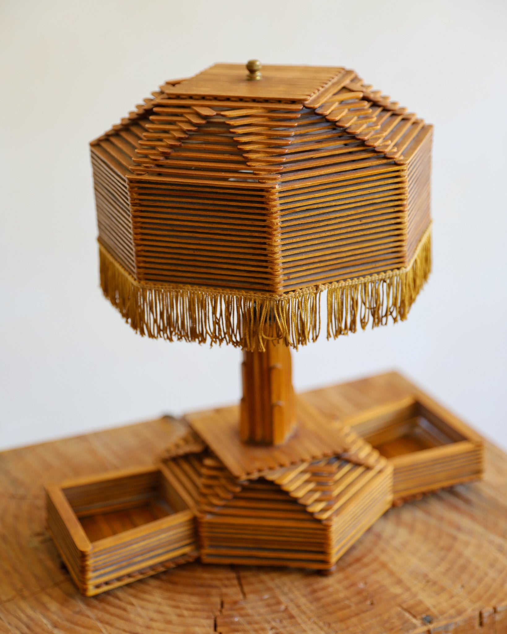 A decorative table lamp made from layered wooden slats, featuring a tiered shade with fringe trim and a geometric base, photographed on a wooden surface against a plain light background.