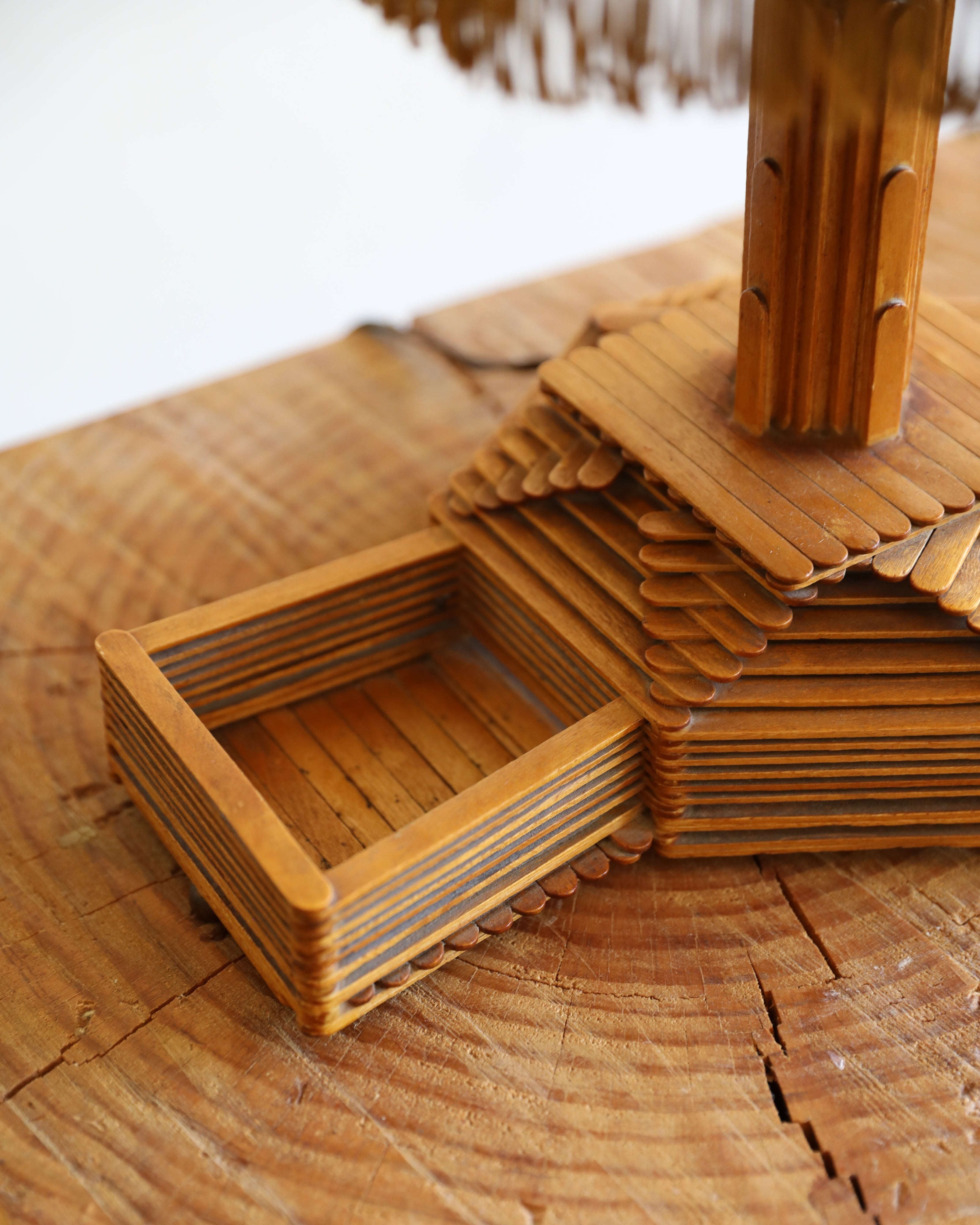 A close-up of a wooden decorative object made from layered popsicle sticks, including a square frame and stacked house-shaped pieces, displayed on a rustic wood surface.
