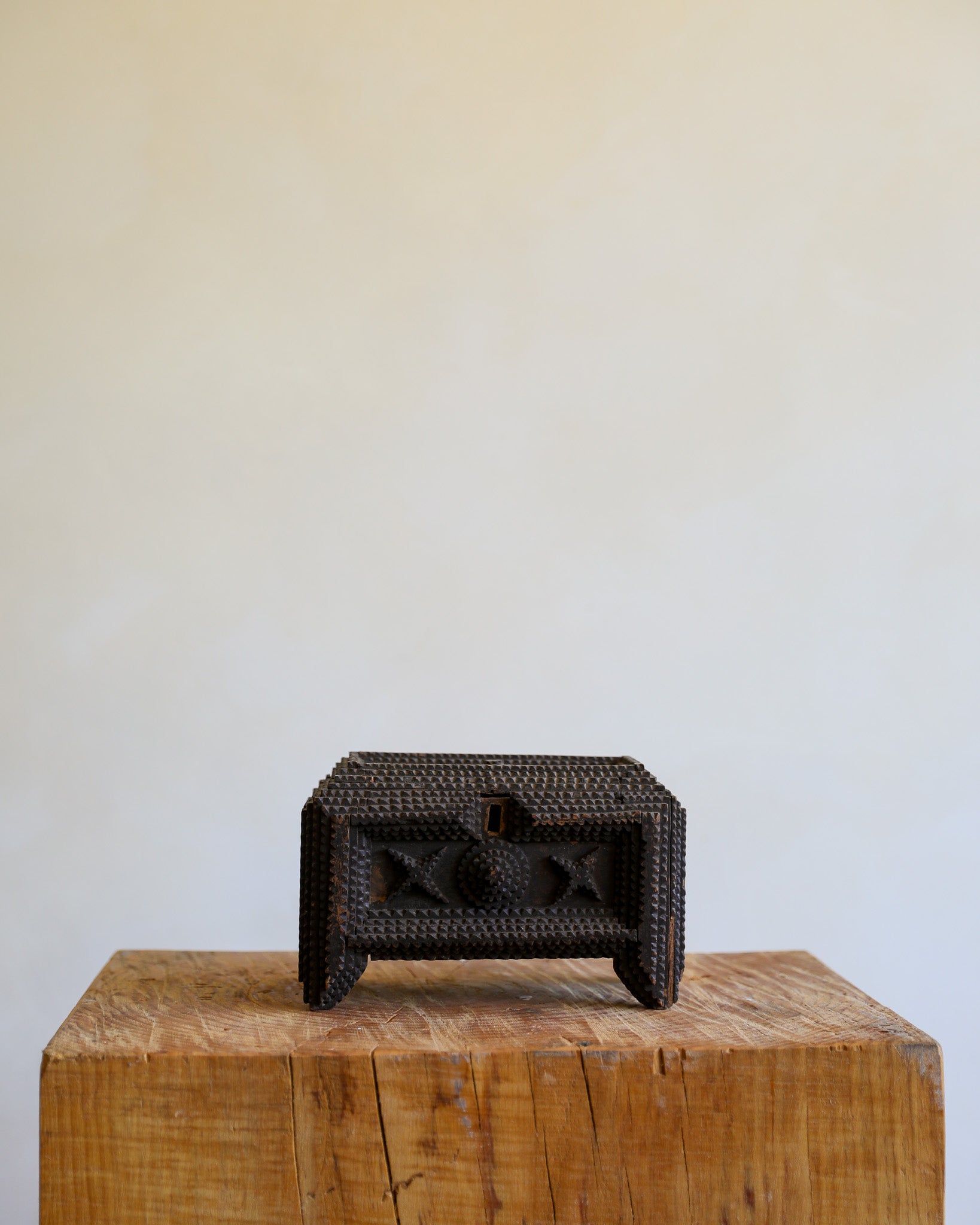 A small dark woven box with a single front drawer and carved geometric details, displayed on a rustic wooden block against a plain light beige wall.