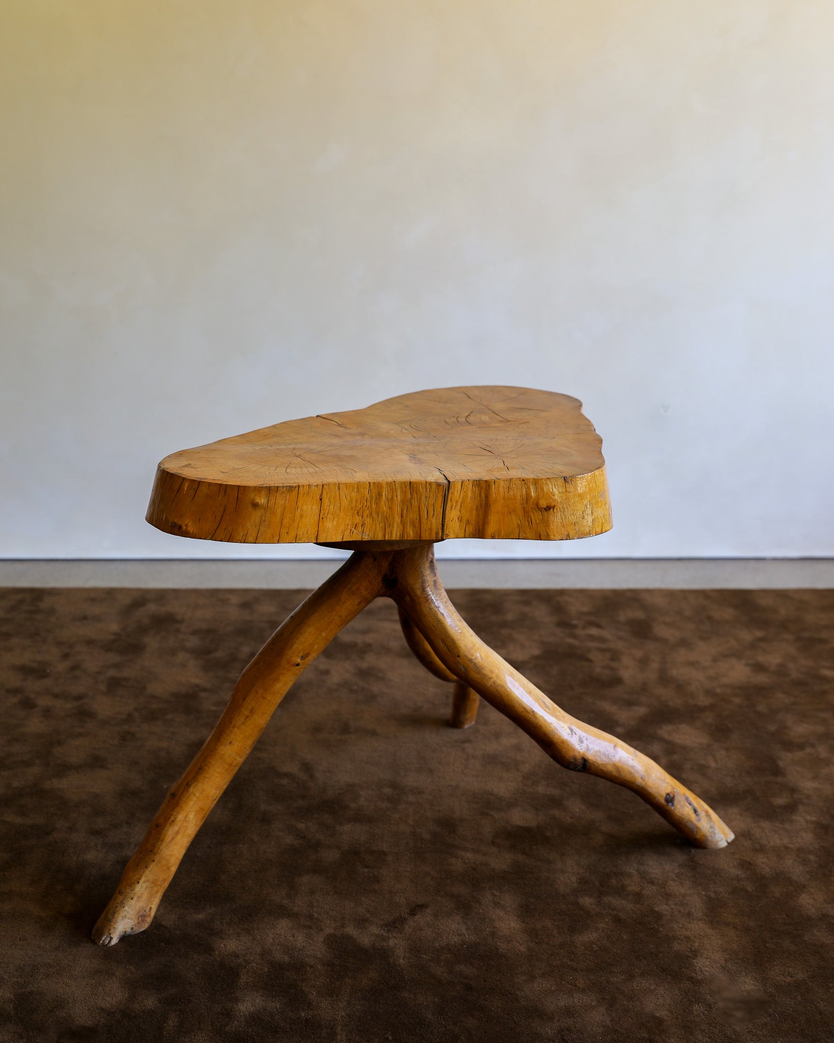 A rustic wooden stool with a thick, irregularly shaped natural tree slice seat and three branching branch-like legs, shown on a dark brown textured floor against a plain light beige wall.