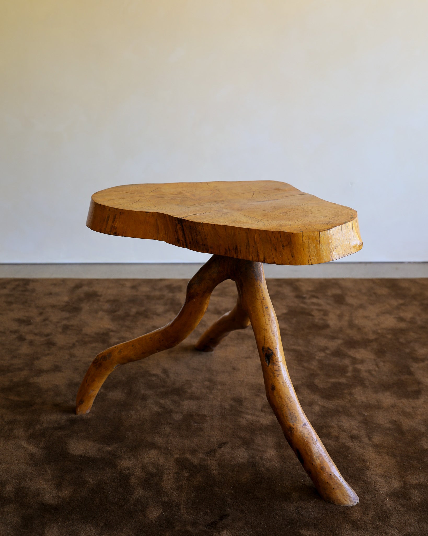 A rustic wooden stool with a natural live-edge seat and three curved branch-like legs, shown on a brown textured rug against a plain light wall.