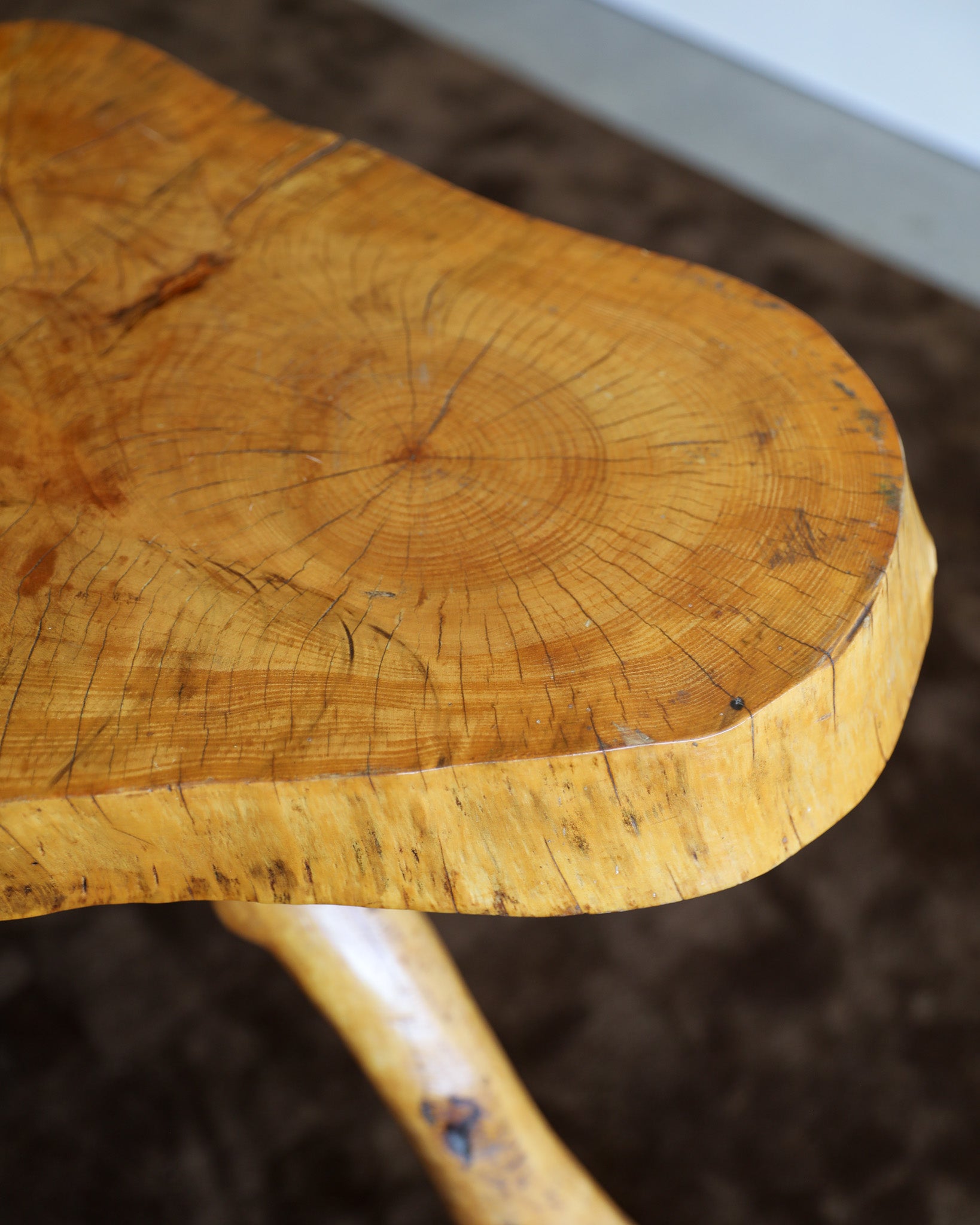 Close-up of a live-edge wooden table top showing natural wood grain, tree growth rings, and a curved live edge, with part of the matching wooden leg visible below against a dark brown floor background.