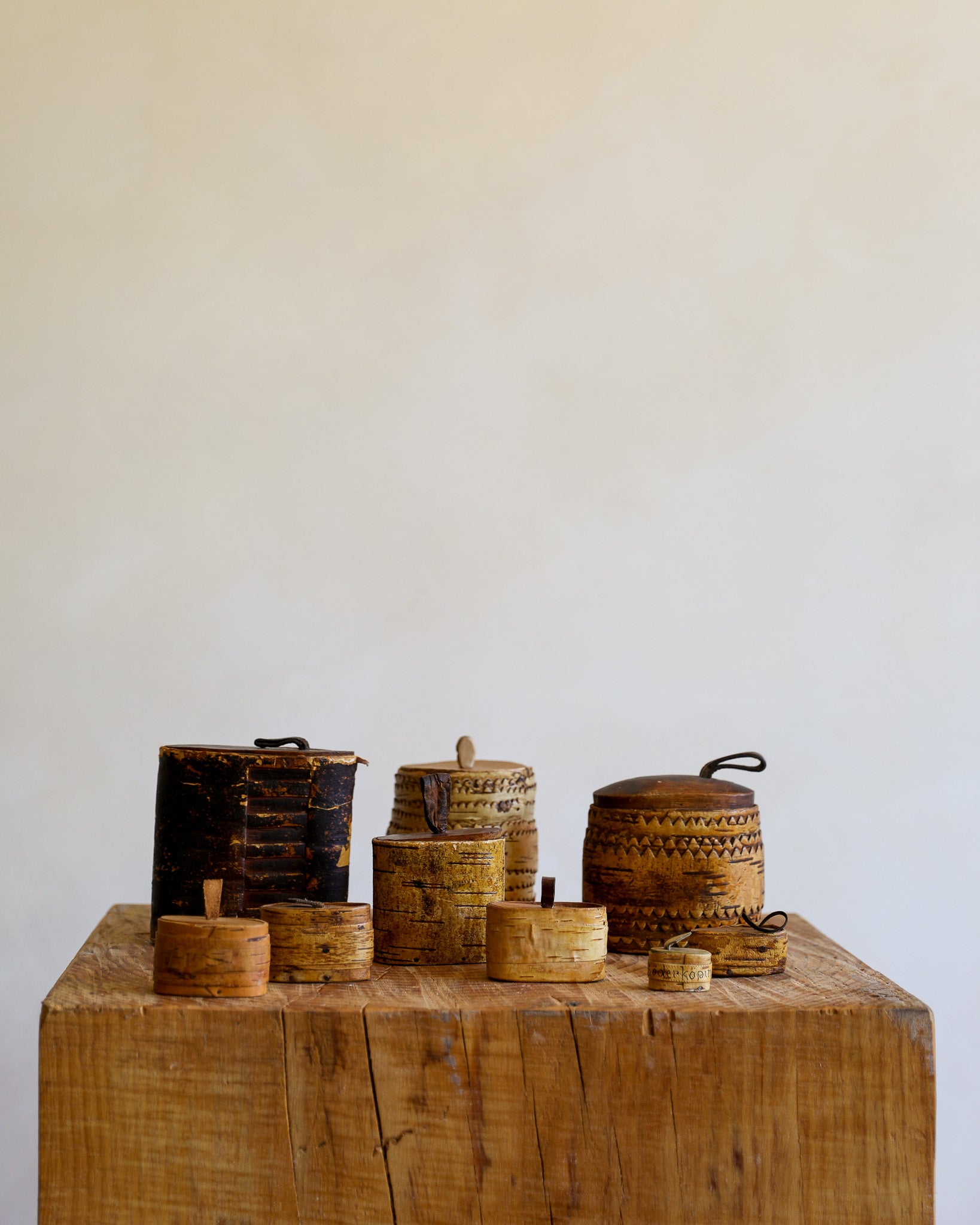 A group of small hand-woven baskets and wooden containers with lids, displayed on a rustic wooden block against a plain off-white wall.