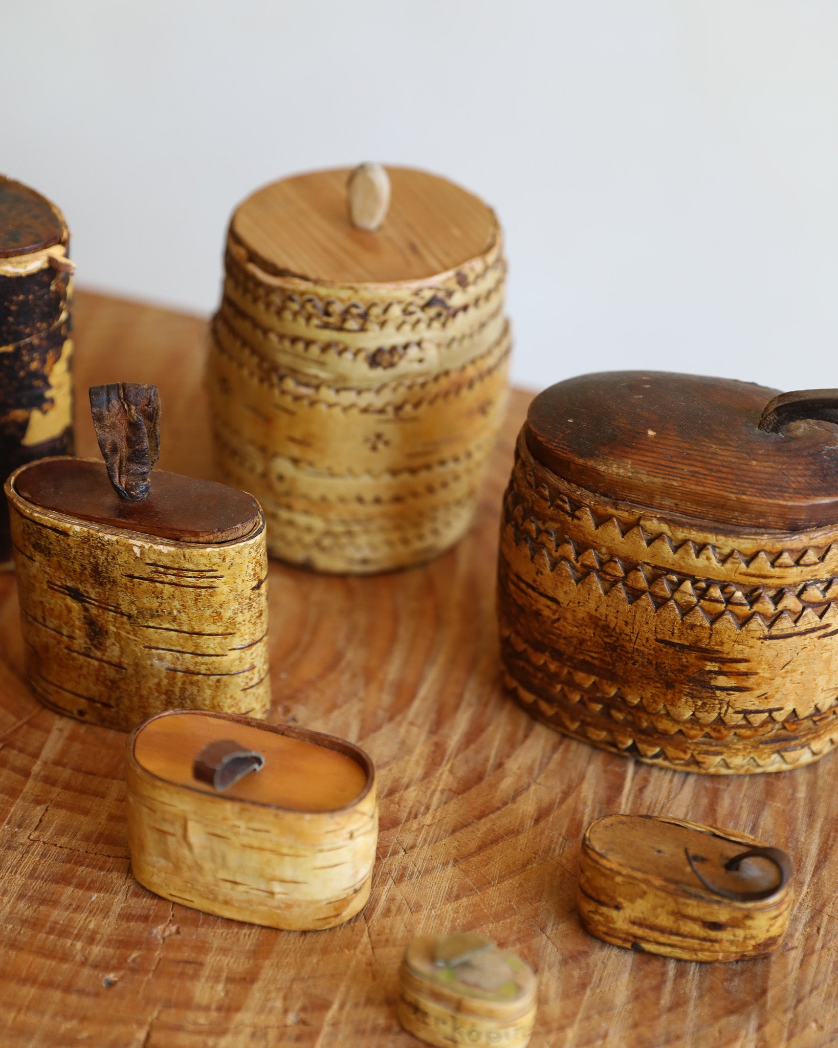 A group of small handcrafted wooden keepsake boxes and containers with natural bark edges, carved geometric patterns, and dark wood lids, arranged on a wooden surface against a light background.