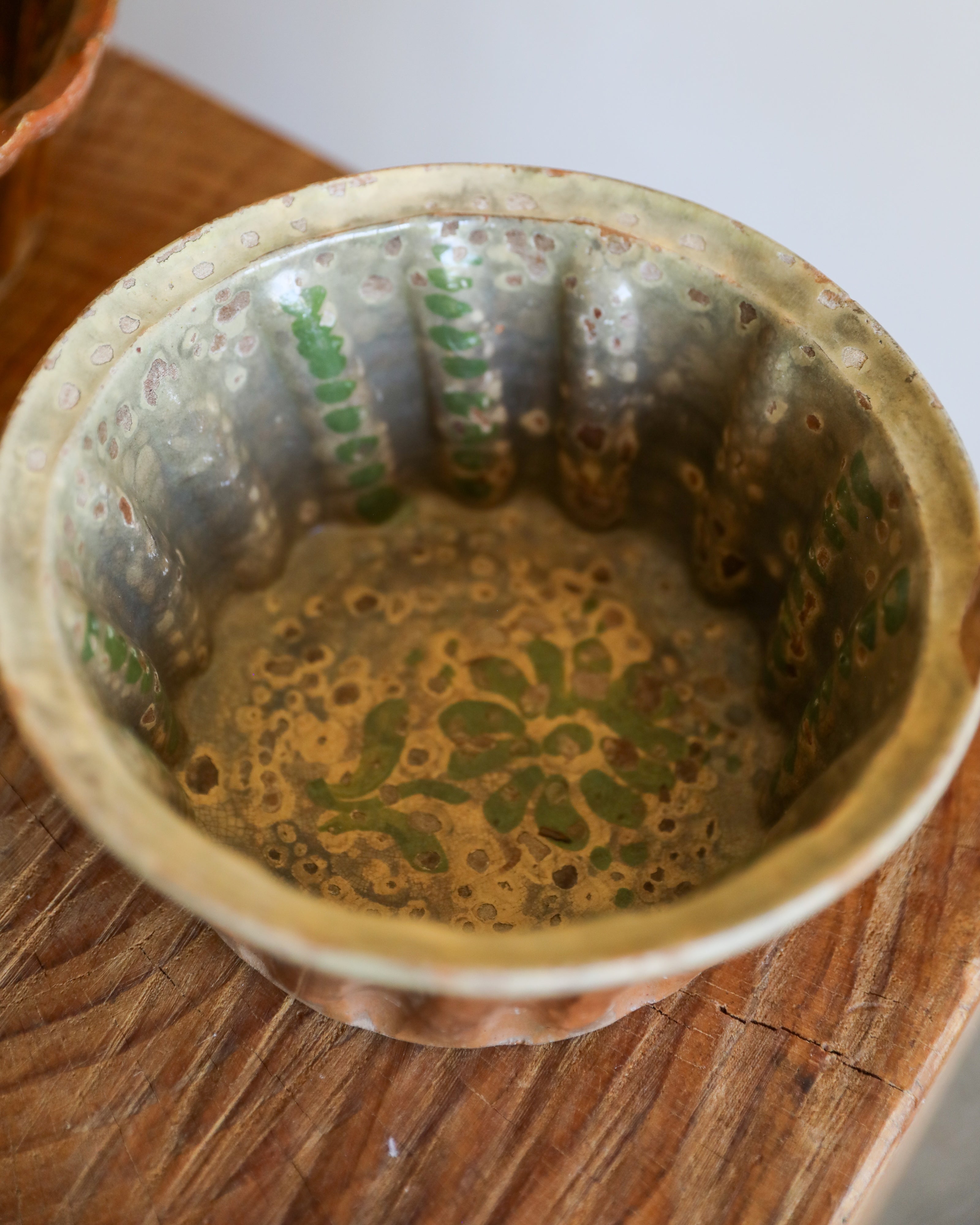 A close-up of a vintage hammered metal bowl with green floral patterns on the inside, resting on a rustic wooden surface.