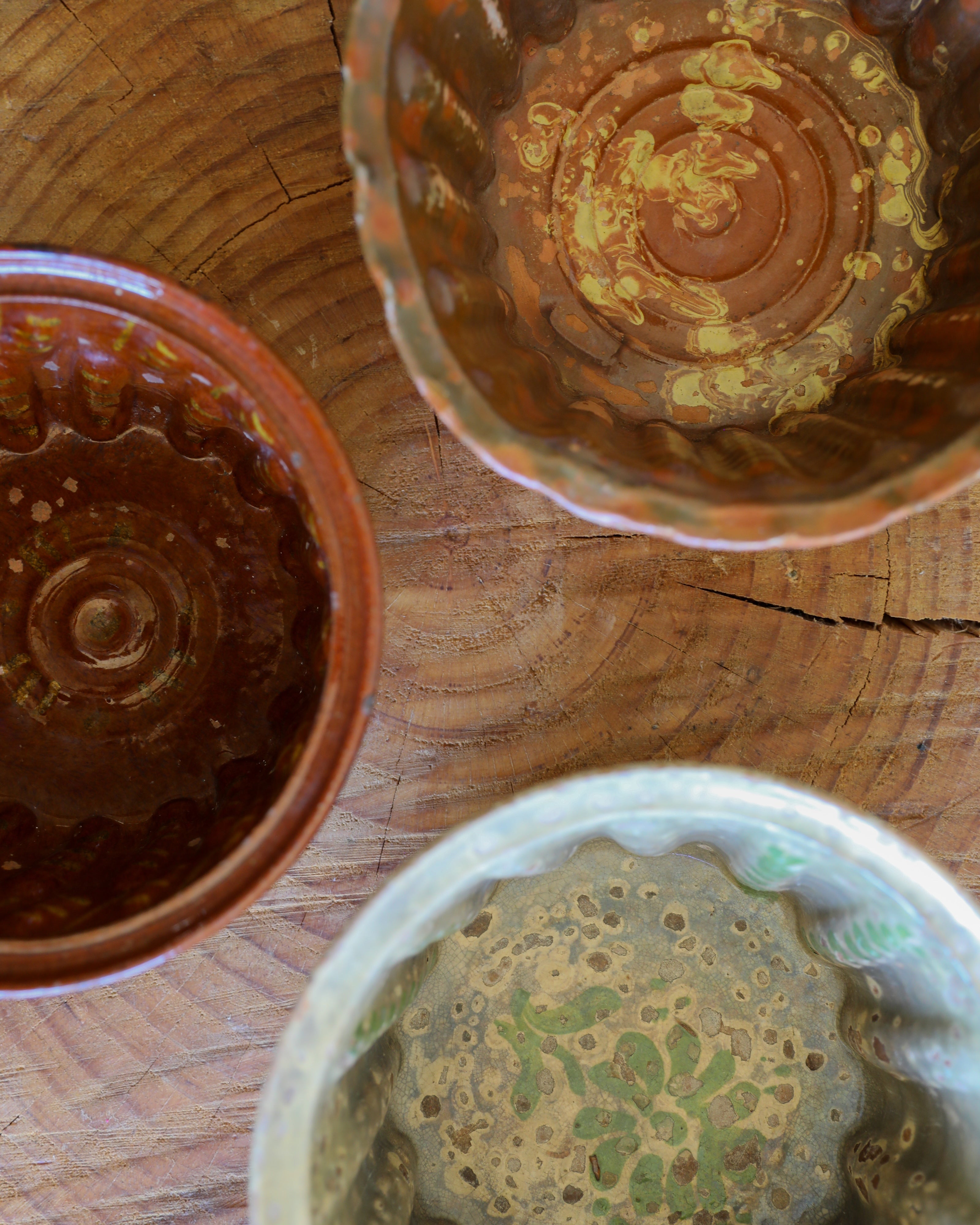 Three handmade ceramic bowls with scalloped rims displayed on a rustic wooden surface. Two bowls are glazed in warm reddish-brown tones, and one is decorated with a textured sage green and beige floral pattern.