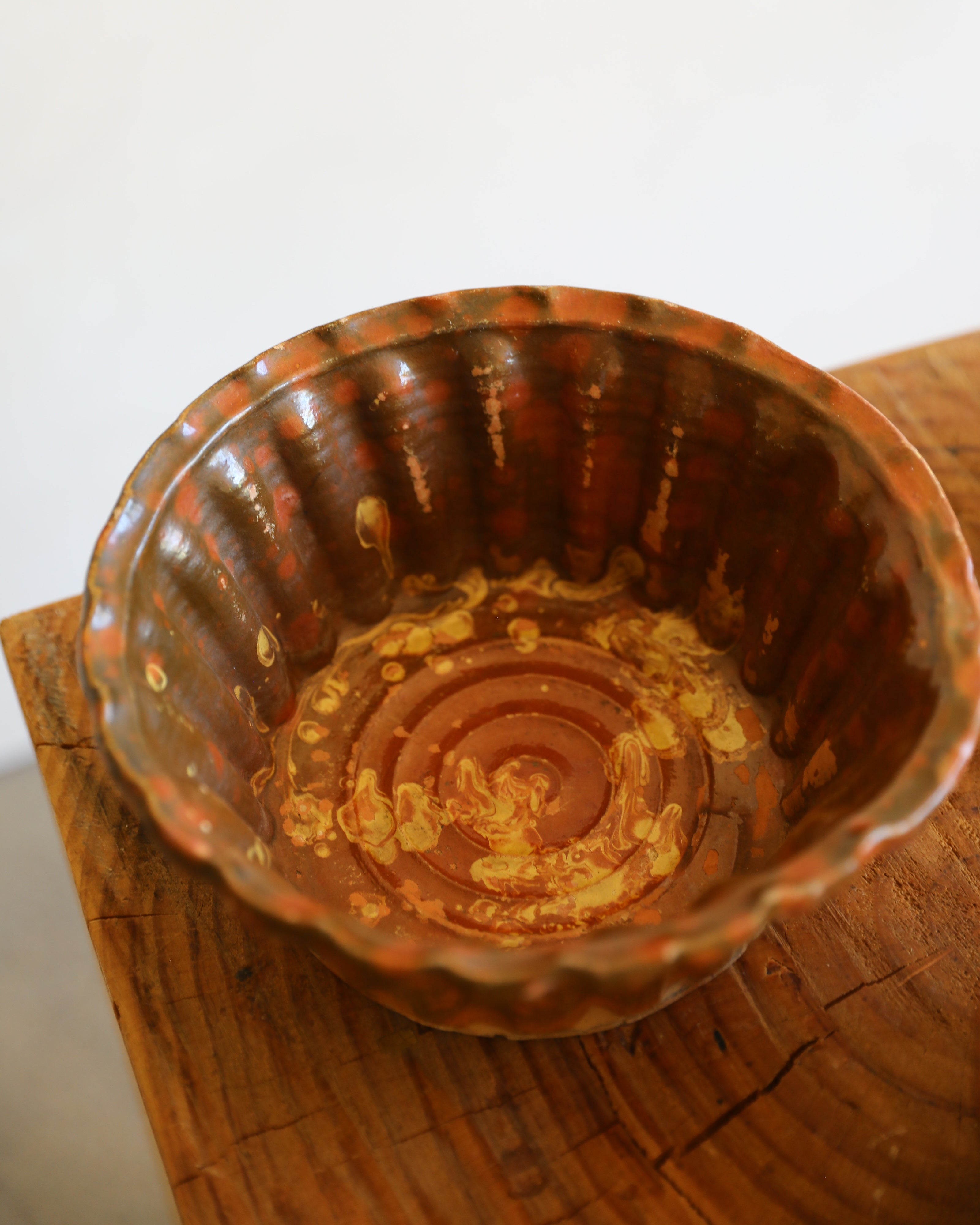 A handmade ceramic bowl with a scalloped rim and a rustic, hand-painted finish in earthy brown and gold tones, resting on a wooden surface against a plain white background.
