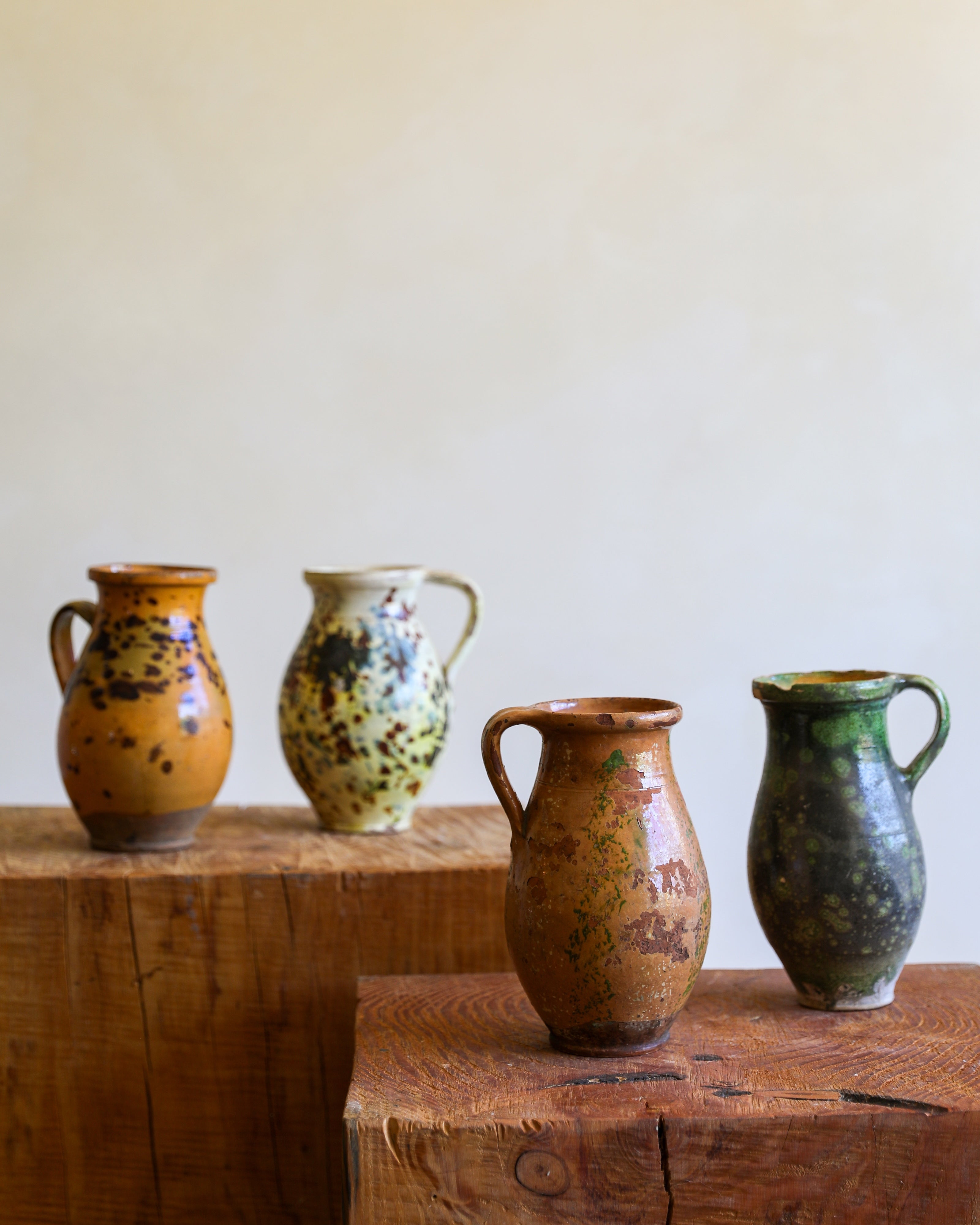 Four handmade ceramic pitchers in various earthy glazes and patterns, displayed on rustic wooden blocks against a plain beige wall.