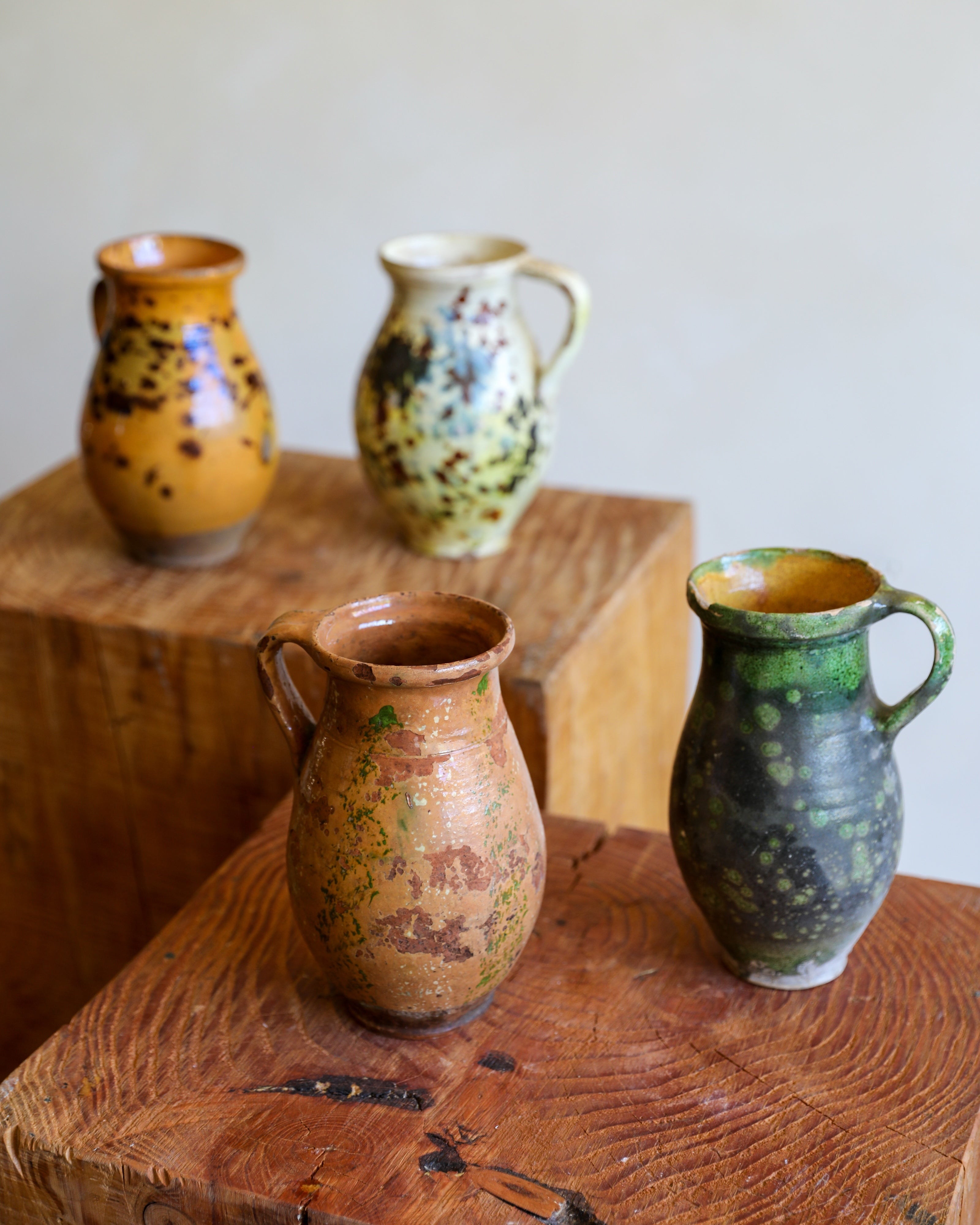 Four handmade ceramic pitchers with rustic glazes displayed on wooden blocks against a neutral background.