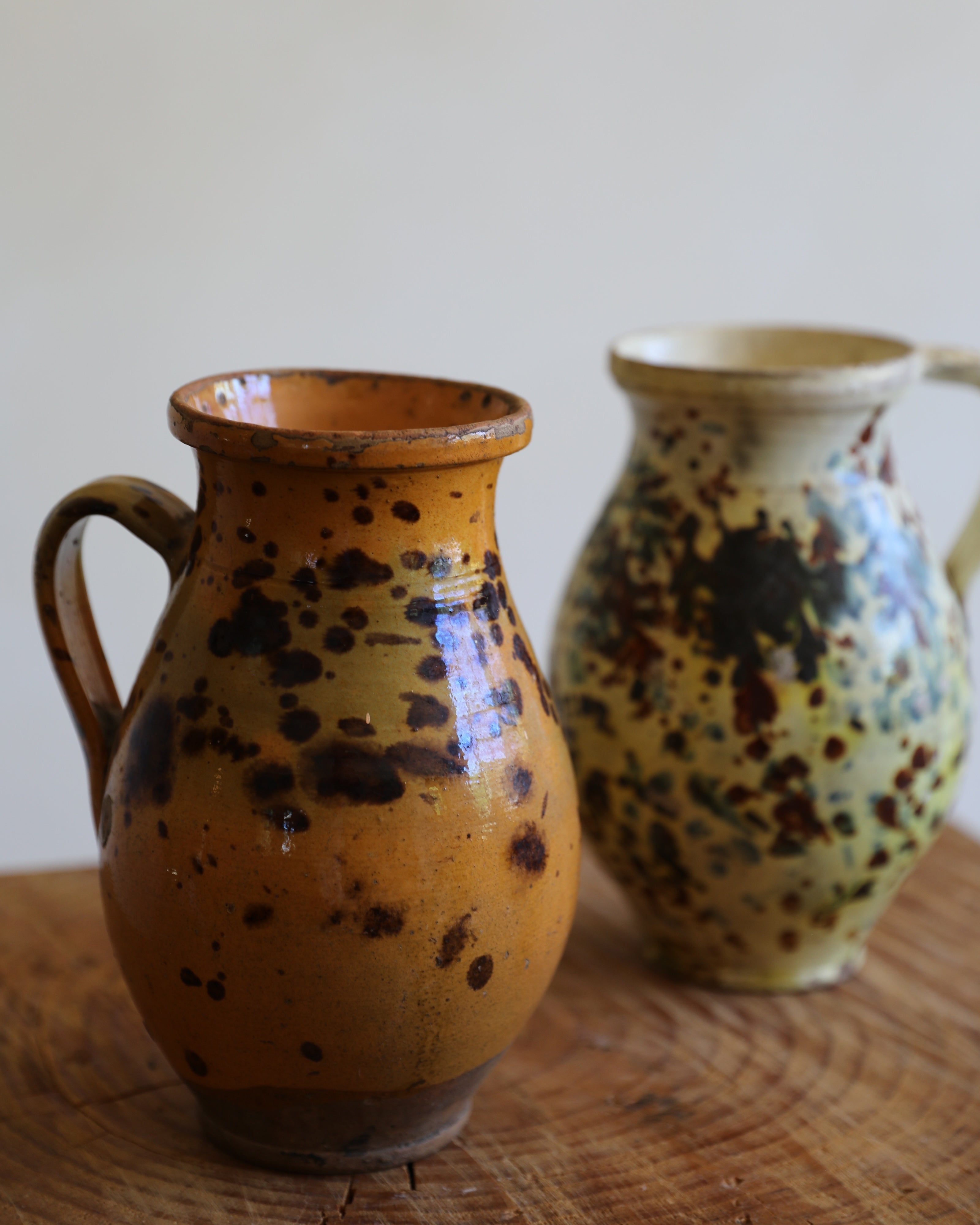 Two handmade ceramic pitchers with speckled glazes, one in the foreground in warm ochre-brown and the other in the background in pale cream with dark brown spots, set on a rustic wooden surface against a neutral off-white wall.