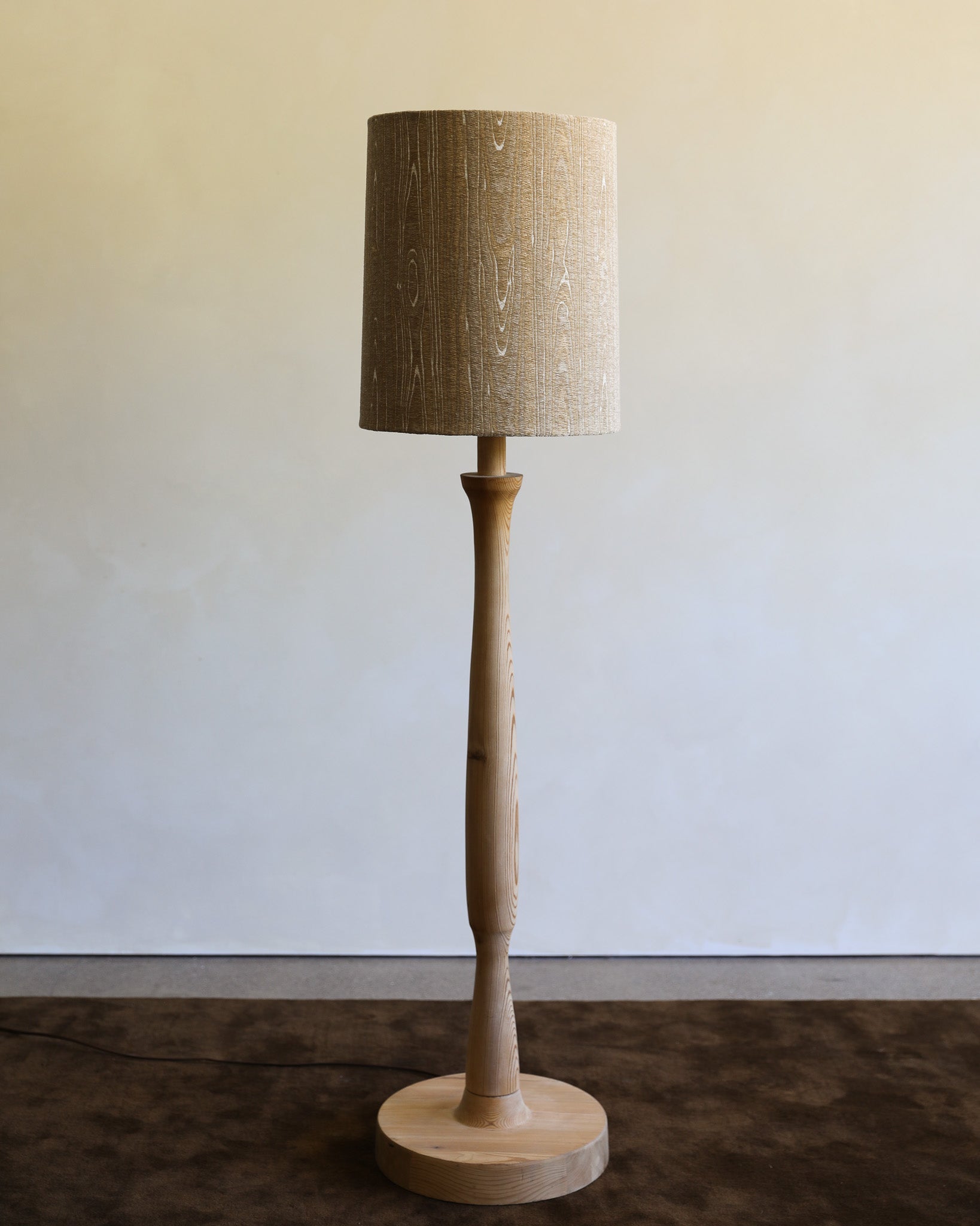 A rustic wooden floor lamp with a tapered drum shade, a sculptural turned wood stem, and a round wooden base, set against a neutral wall.