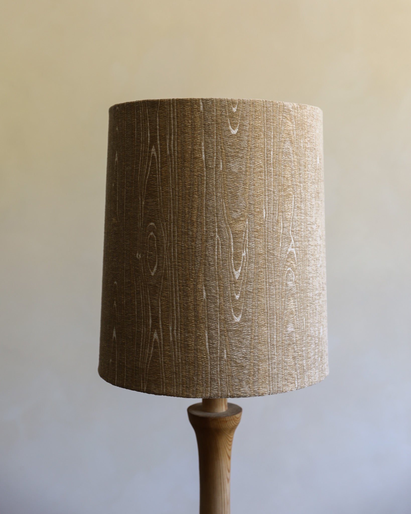 A cylindrical table lamp shade made of textured beige linen fabric with a vertical wood grain pattern, mounted on a wooden lamp base against a neutral beige background.