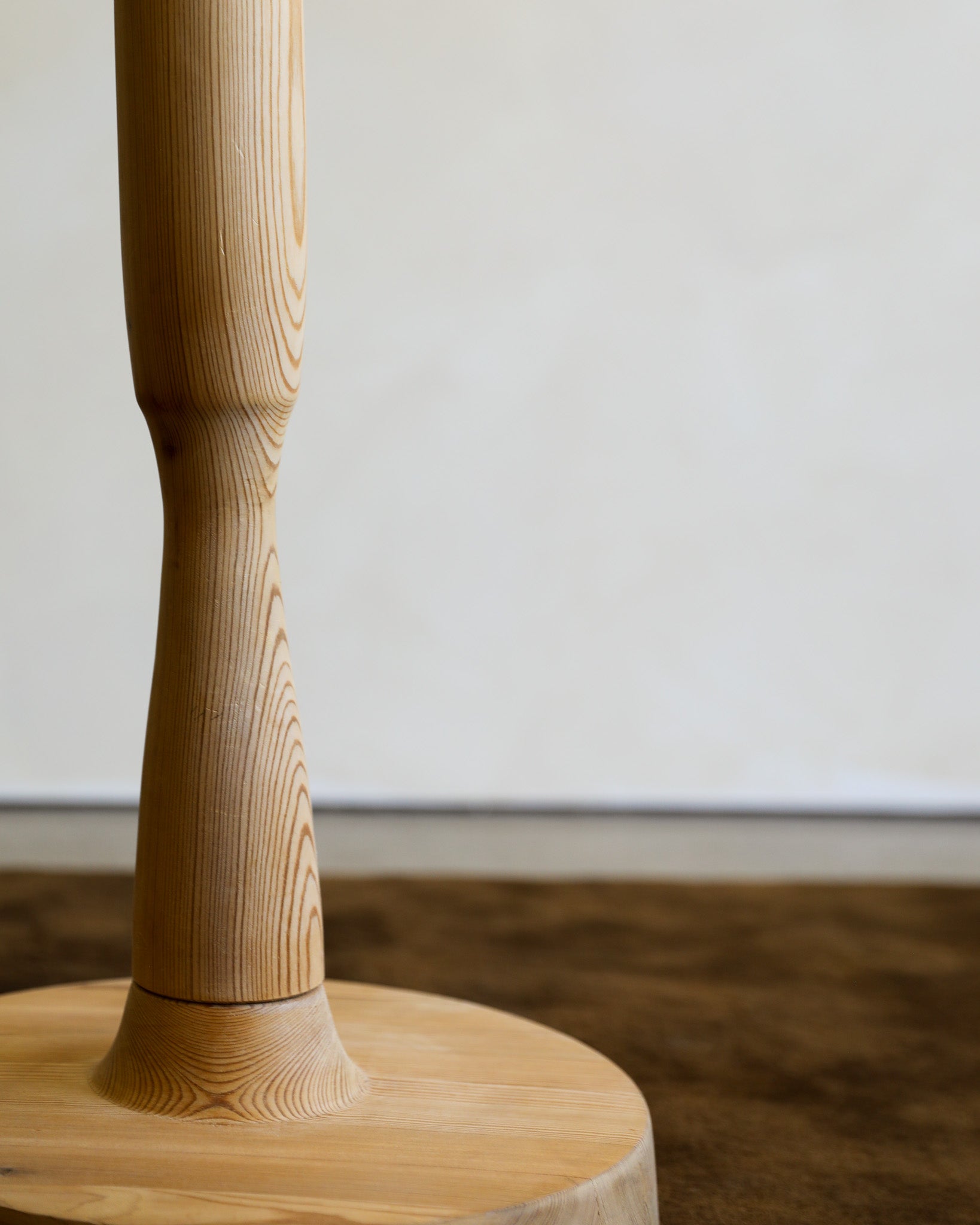 Close-up of a light natural wood furniture leg mounted on a round wooden base, with a plain off-white wall and dark brown floor in the background.