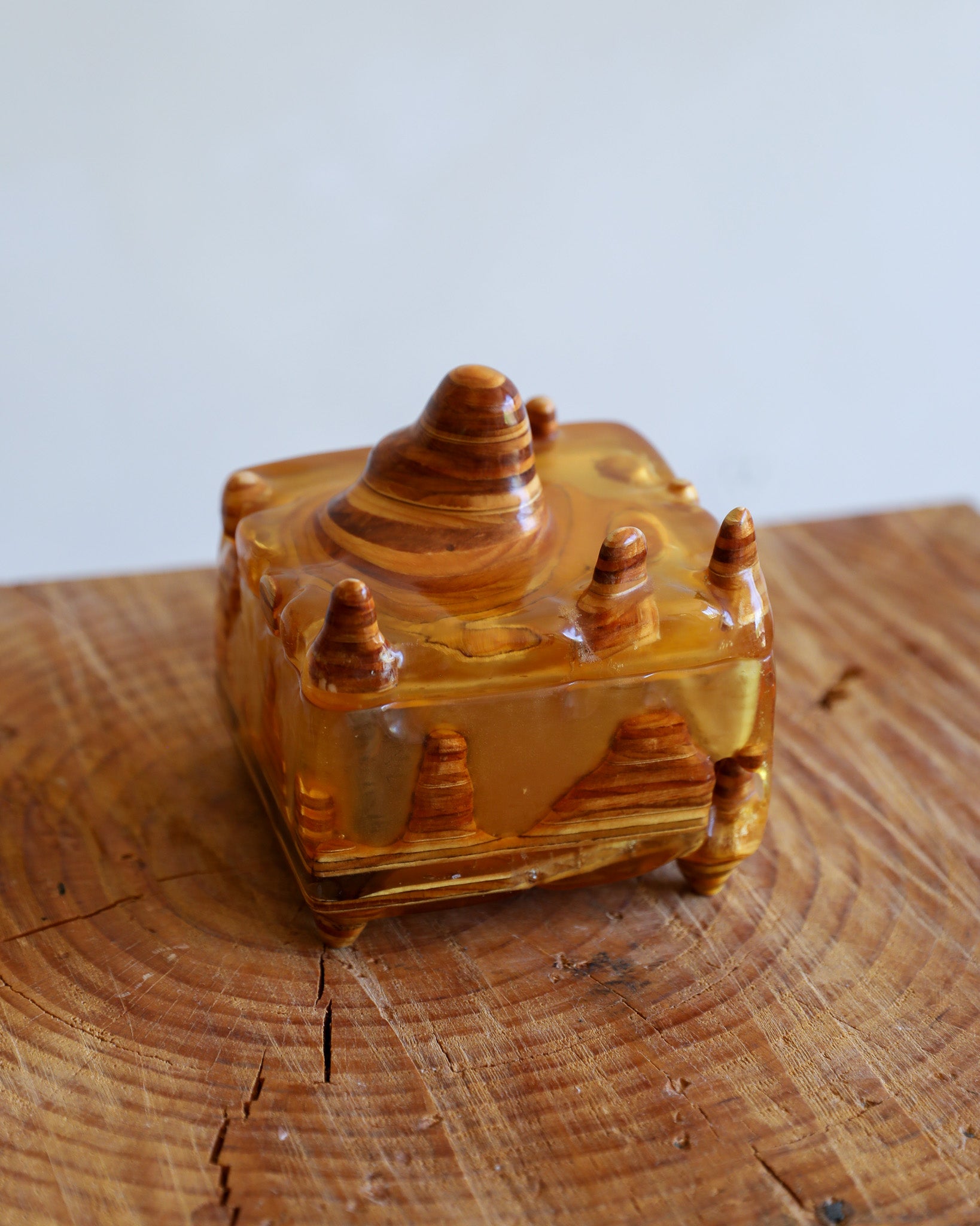 A small carved amber or resin box with a lid, featuring a desert canyon and pine tree motif, displayed on a rustic wood slice against a light gray background.