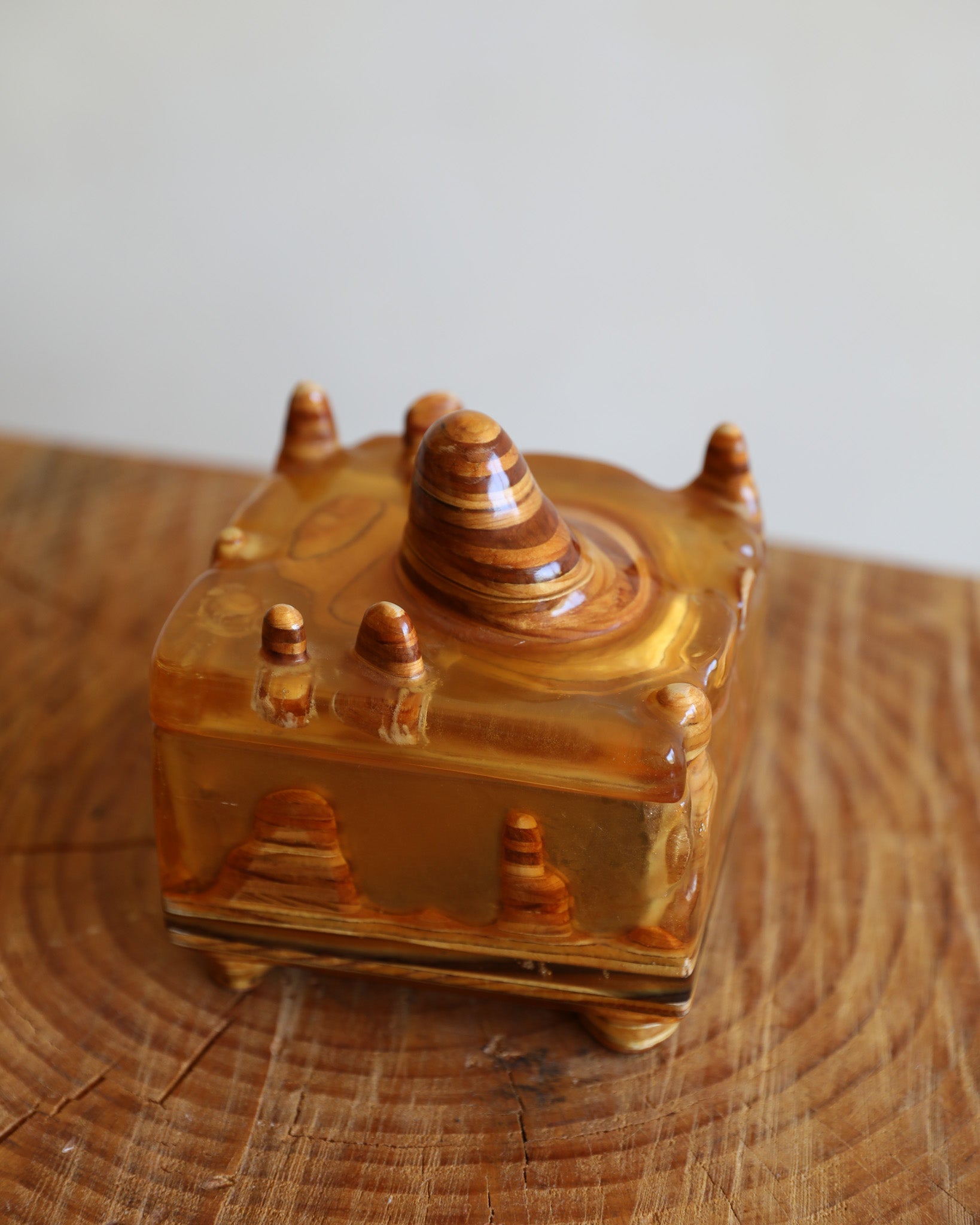A small carved amber resin box with a lid, featuring a central dome and tiered side spires, displayed on a rustic wooden surface against a light gray background.