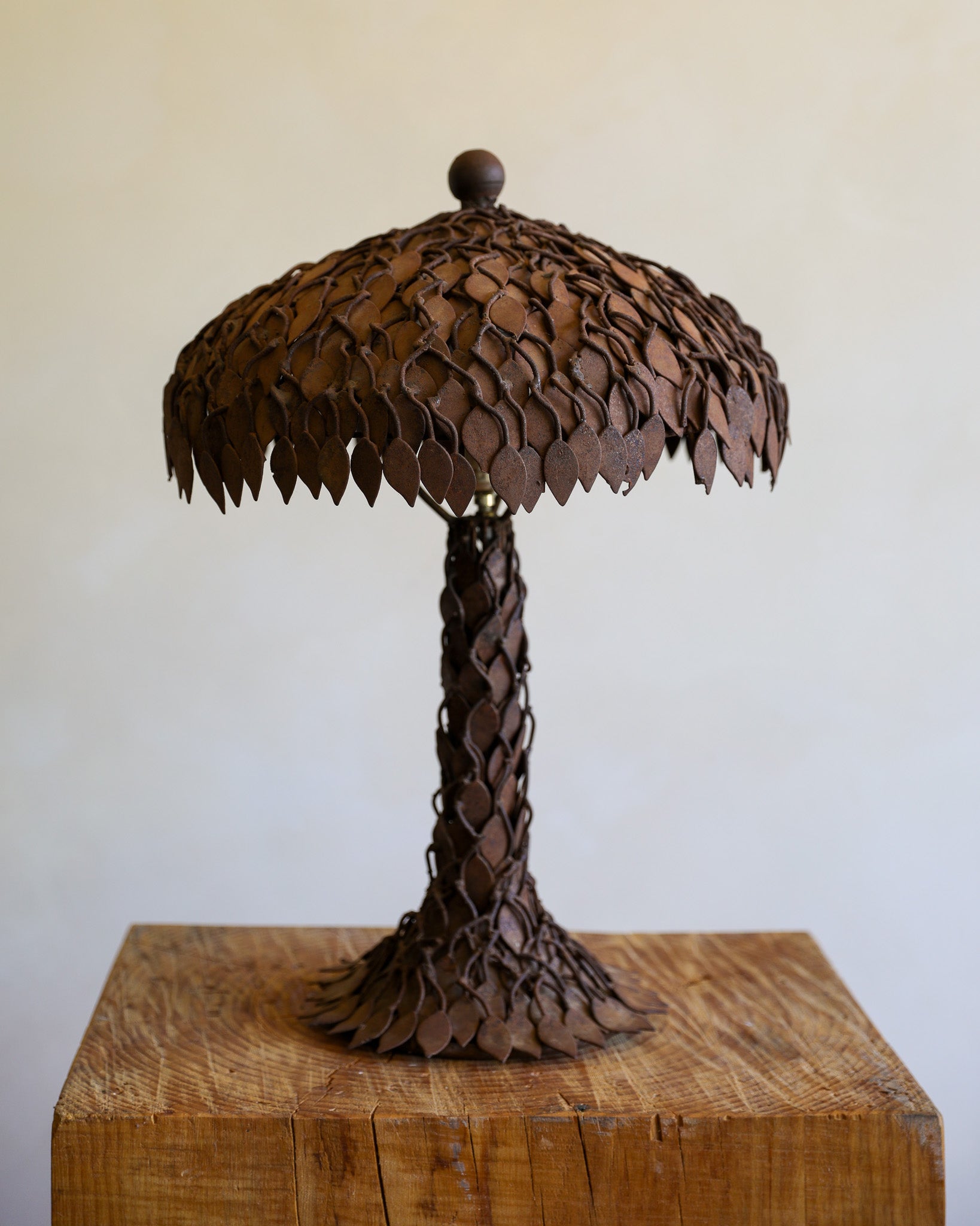 A rustic metal sculpture of a palm tree with a textured, leaf-like crown and trunk, displayed on a wooden block against a plain beige background.