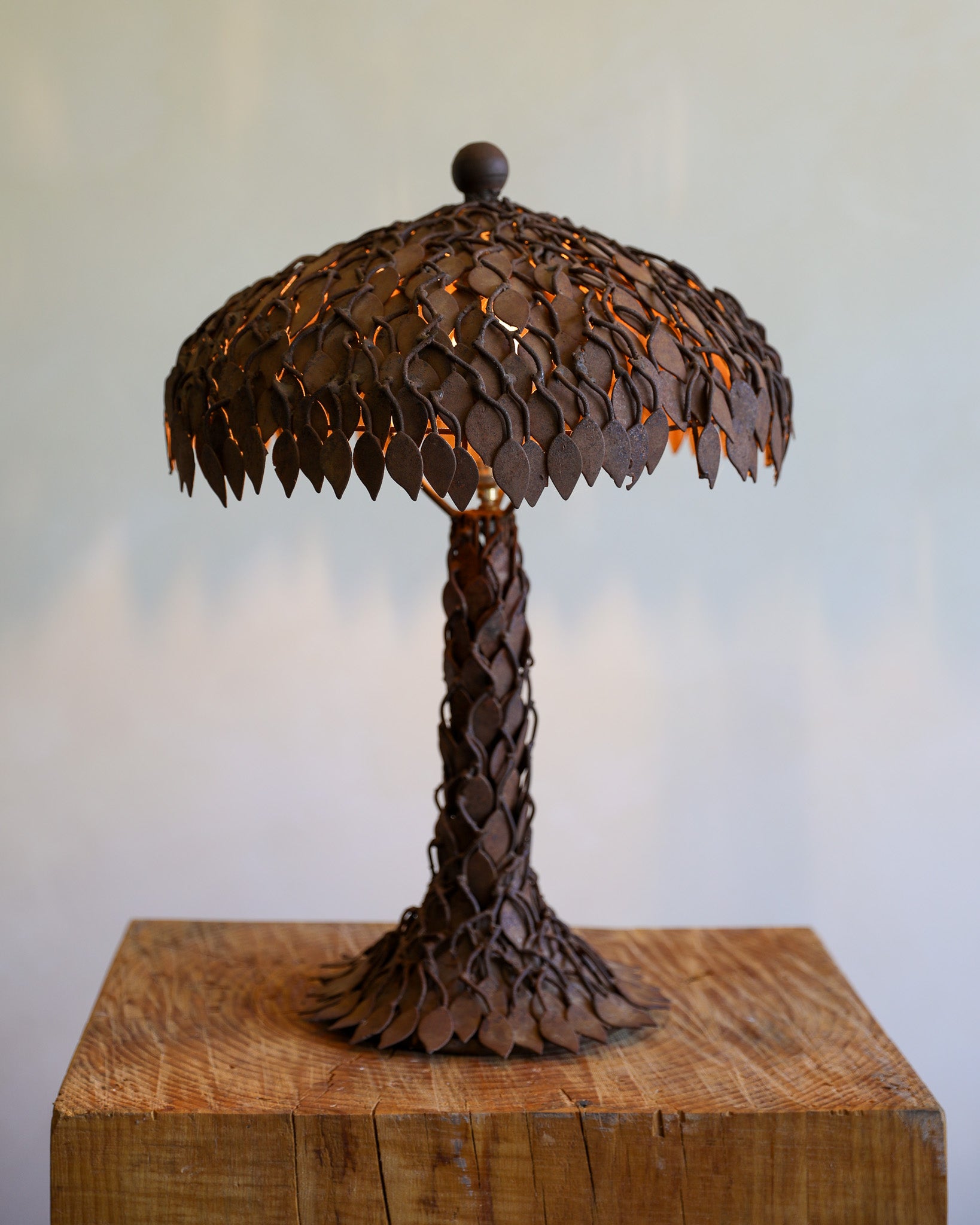 A rustic metal table lamp shaped like a palm tree, featuring a textured shade and trunk made of overlapping leaf-shaped metal cutouts, illuminated from within, set on a wooden block against a soft light gray background.