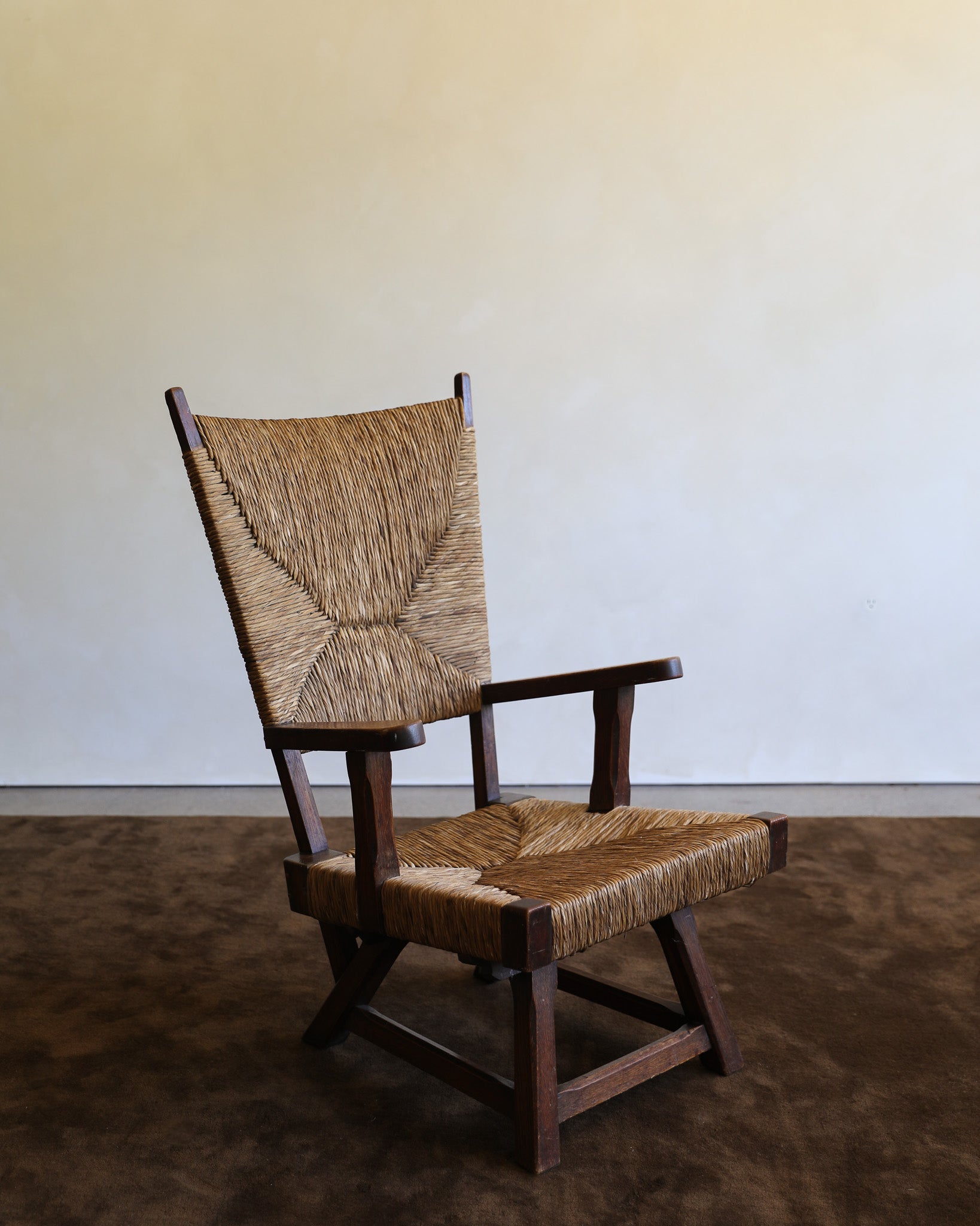 A mid-century modern wooden armchair with a woven natural-fiber seat and backrest, set against a plain beige wall and dark brown floor.