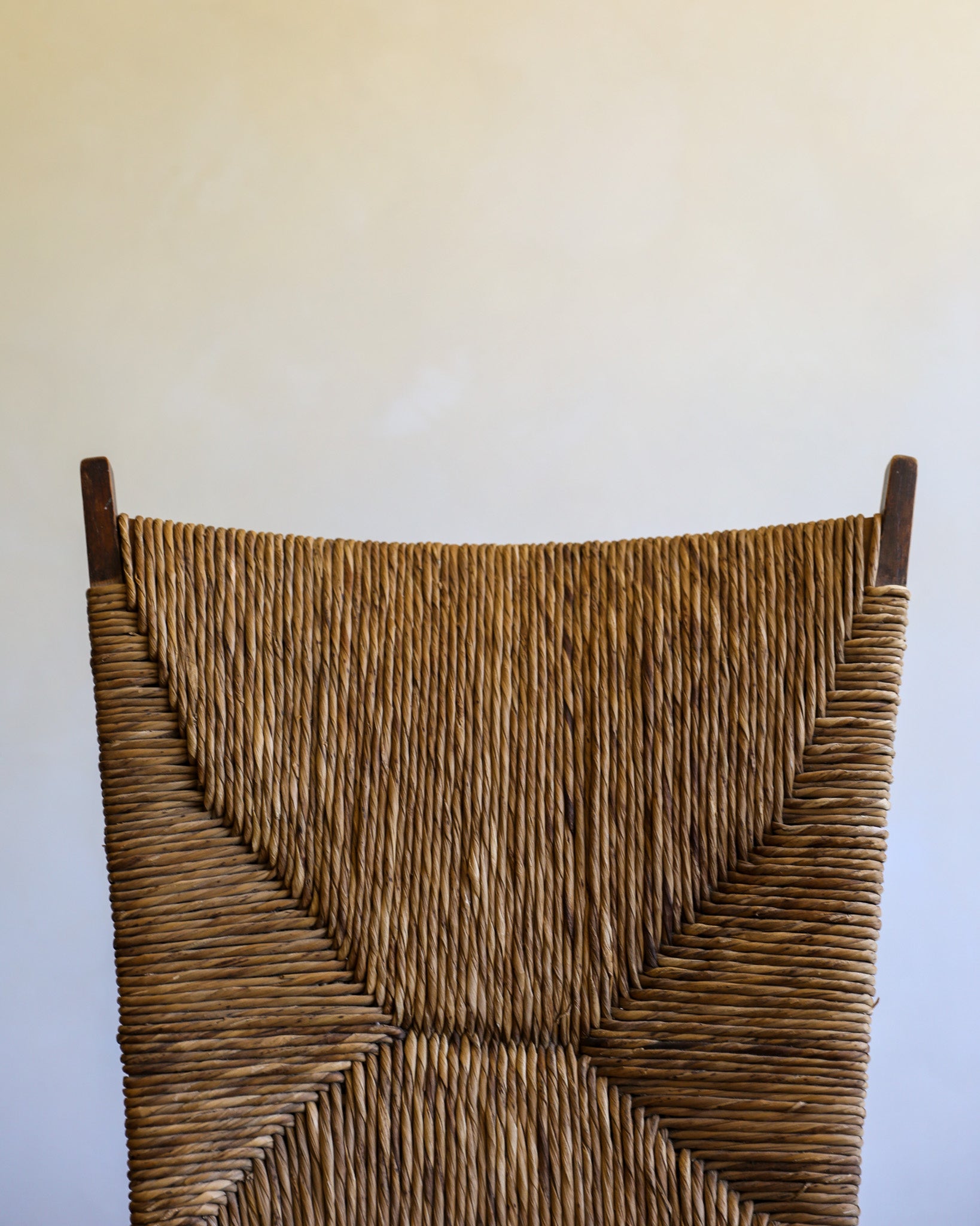 Close-up of a woven natural-fiber chair back with dark wooden finials at the top, shown against a plain off-white wall.