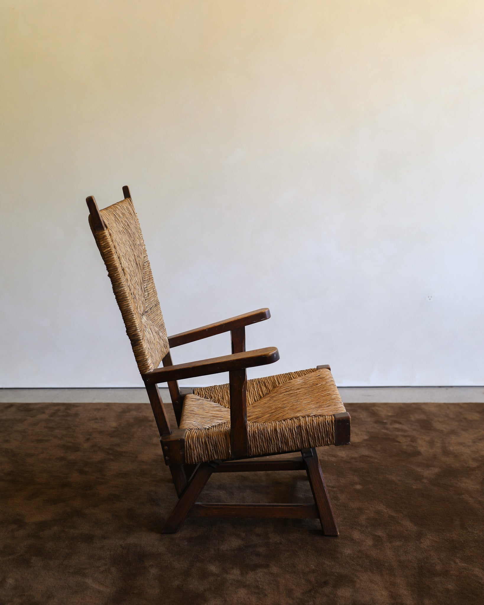 A mid-century modern wooden armchair with a high back and woven natural-fiber seat and back, shown in profile against a plain off-white wall and brown textured floor.