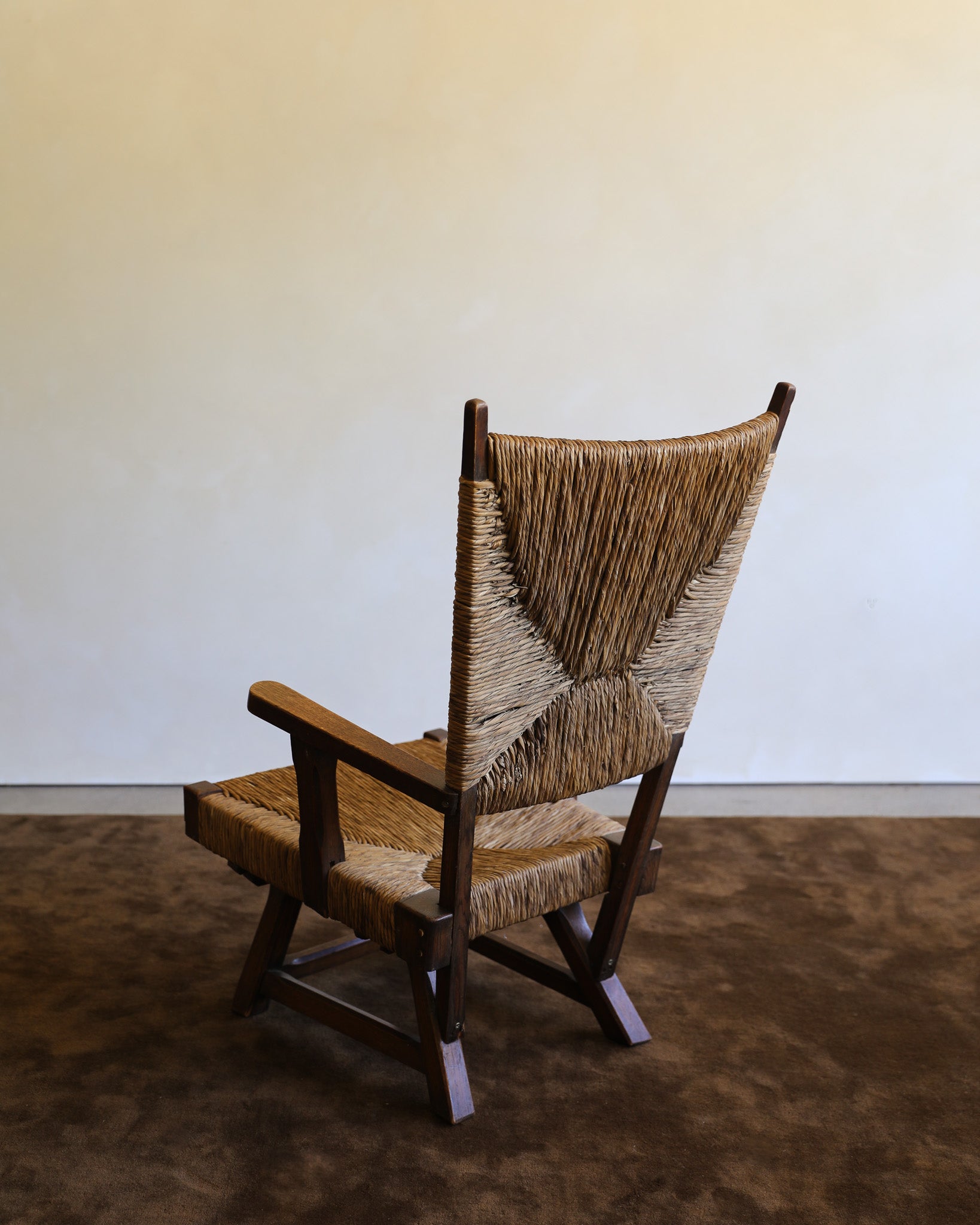 A mid-century modern wooden armchair with woven natural-fiber seats and back, photographed in a minimalist room with a plain wall and textured brown floor.