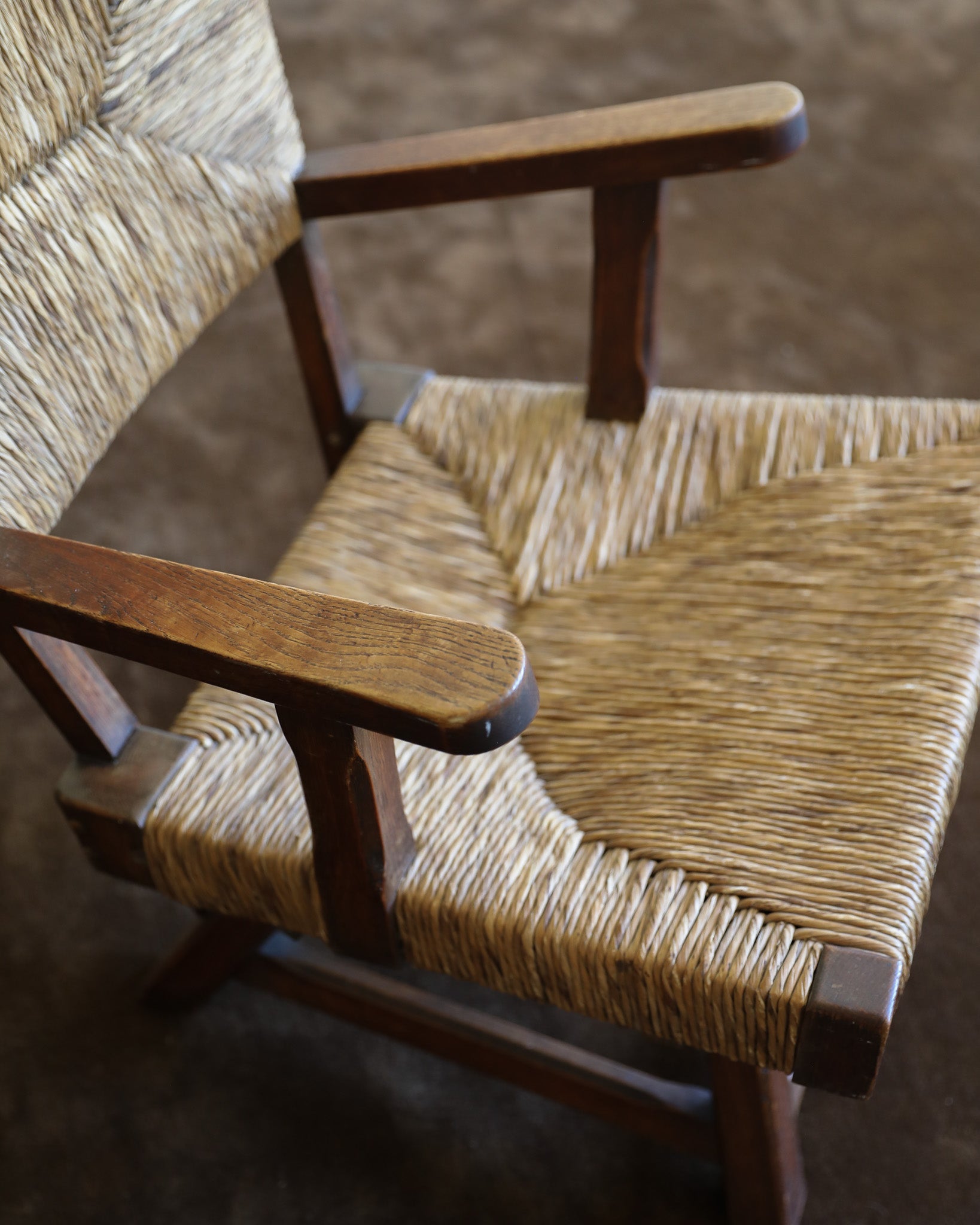 A close-up of a wooden armchair with woven natural-fiber seats and back, shown on a brown textured floor.