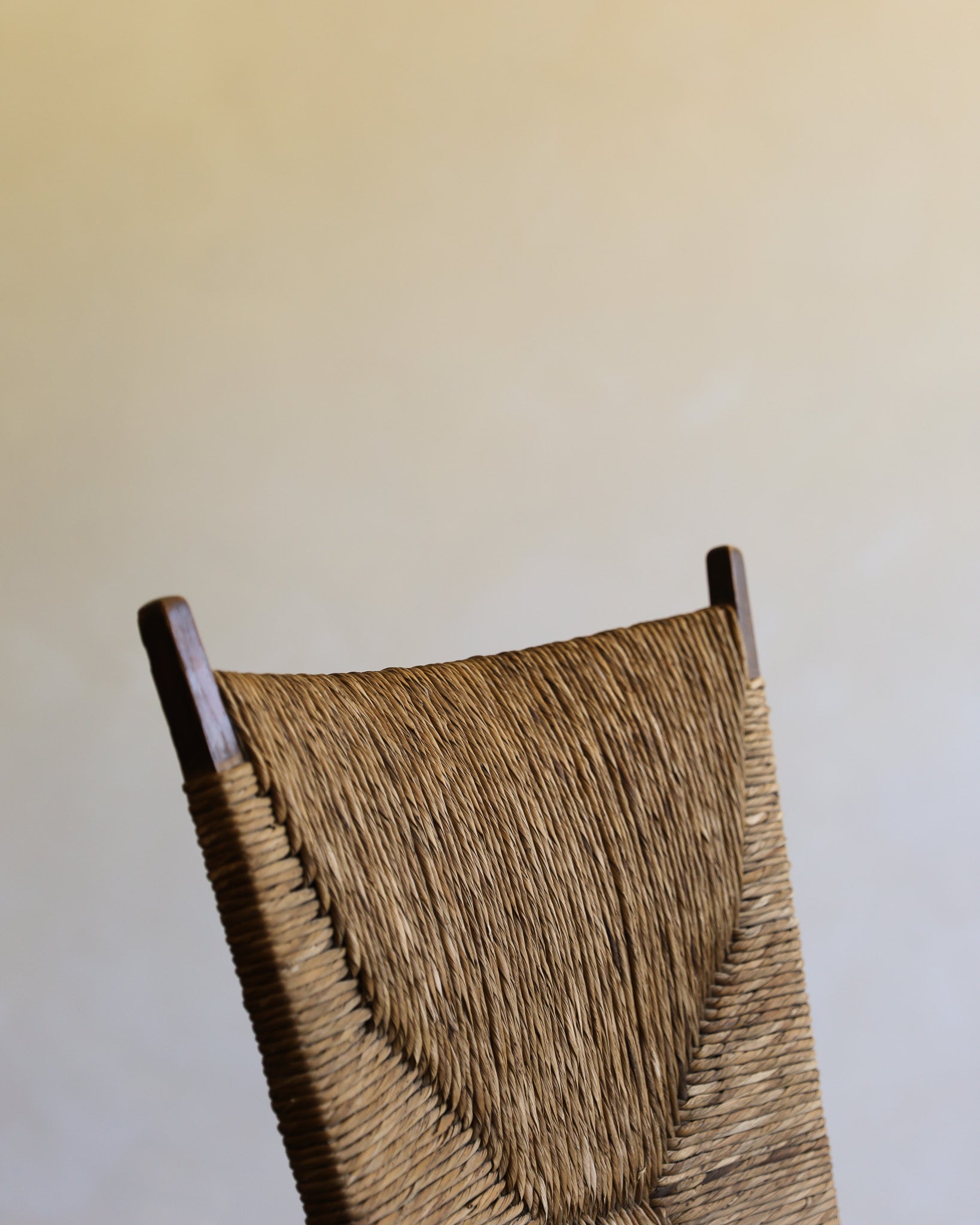 Close-up of a woven chair back with a natural straw or raffia texture and dark wooden finials, set against a plain light beige wall.