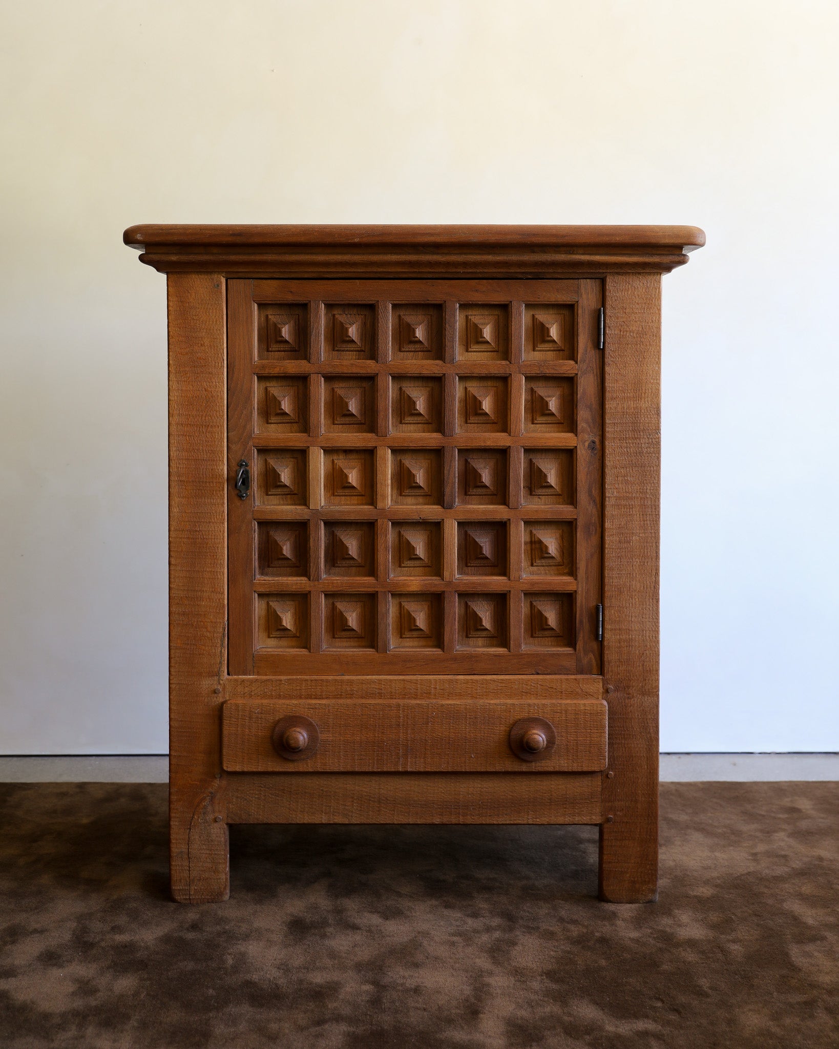 A small wooden cabinet with a carved grid pattern on the door and a single drawer below, shown against a plain light wall and brown rug.