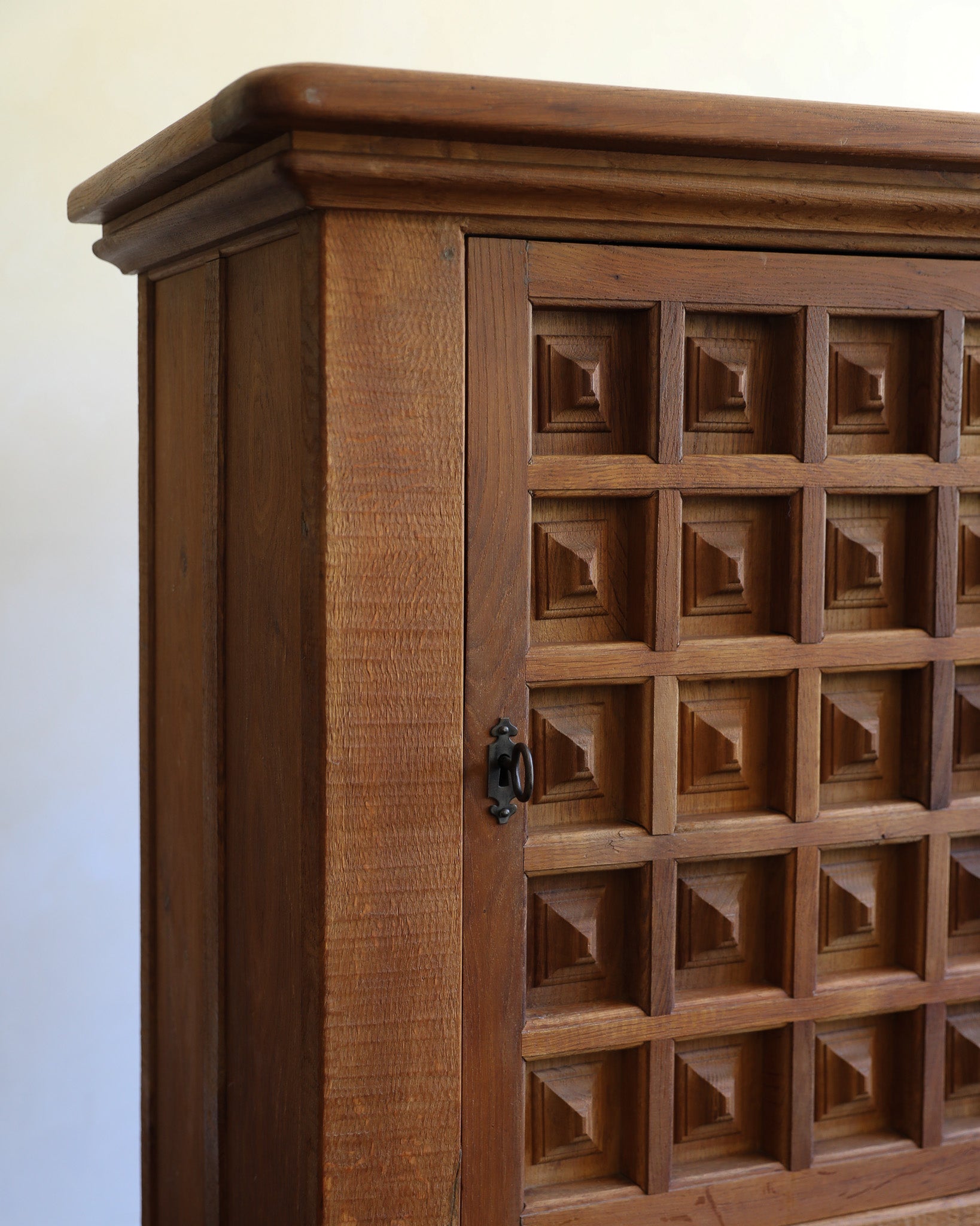 Close-up of a medium brown wooden cabinet with a carved geometric patterned door and a small black metal handle, set against a plain off-white wall.