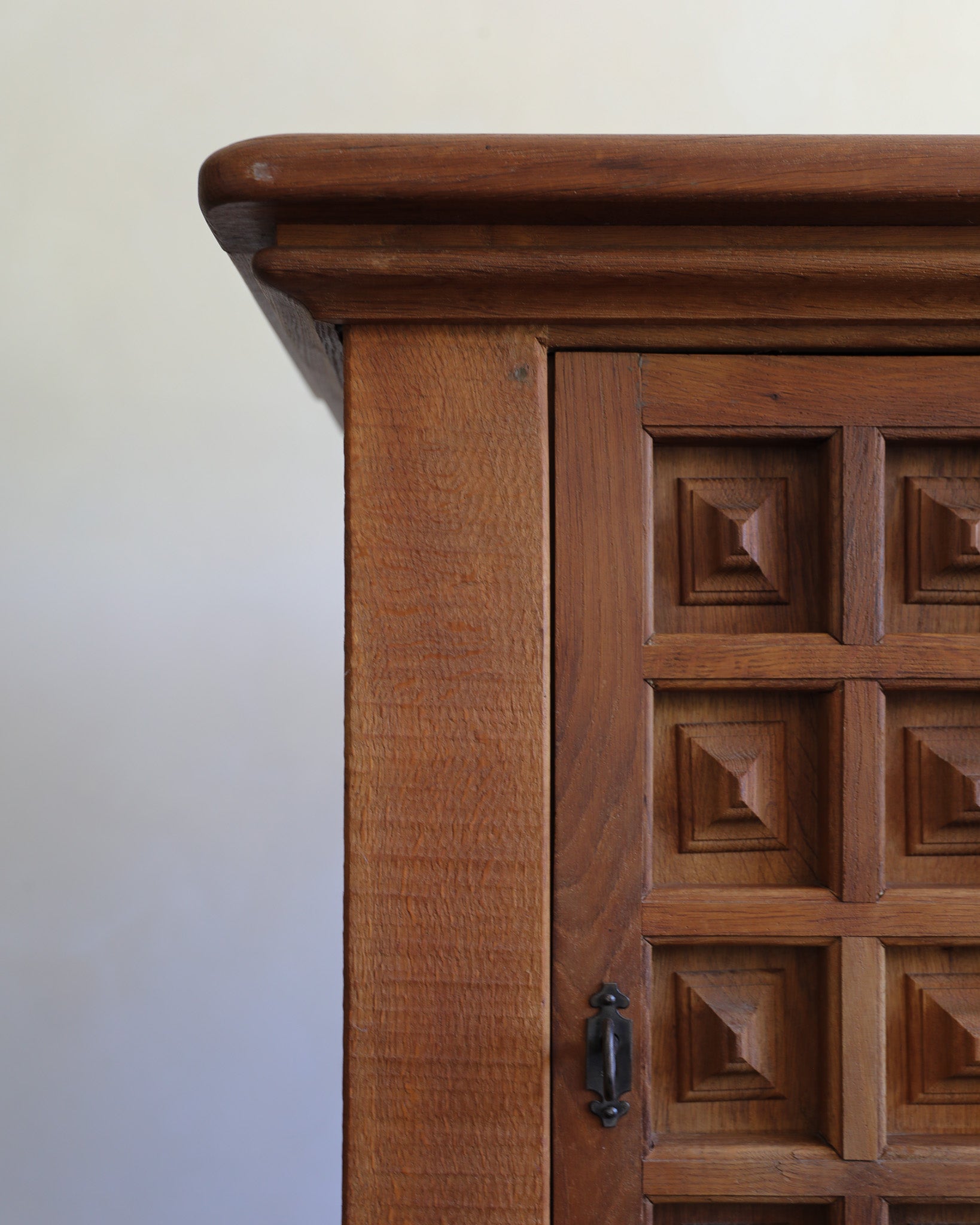 Close-up of a warm medium-brown wooden cabinet corner with carved geometric paneling on the door and a dark metal handle, set against a plain off-white wall.
