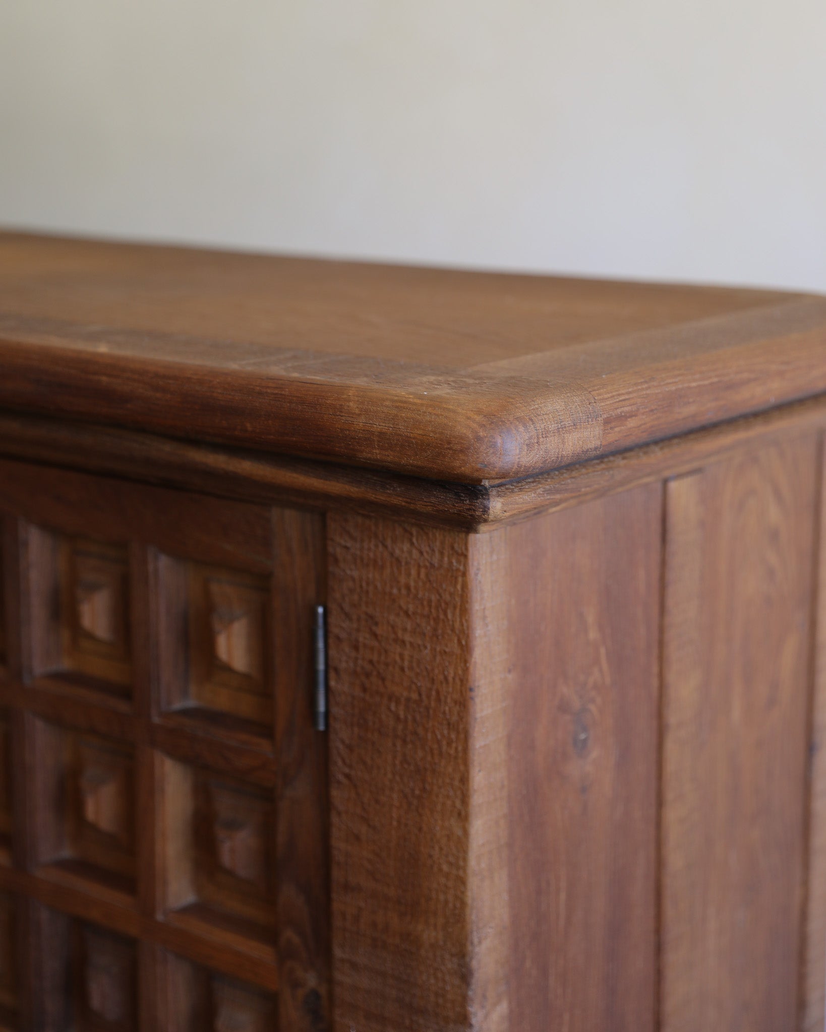 Close-up of a dark brown wooden cabinet corner showing the top surface, carved door panel, side edge, and wood grain against a plain light background.