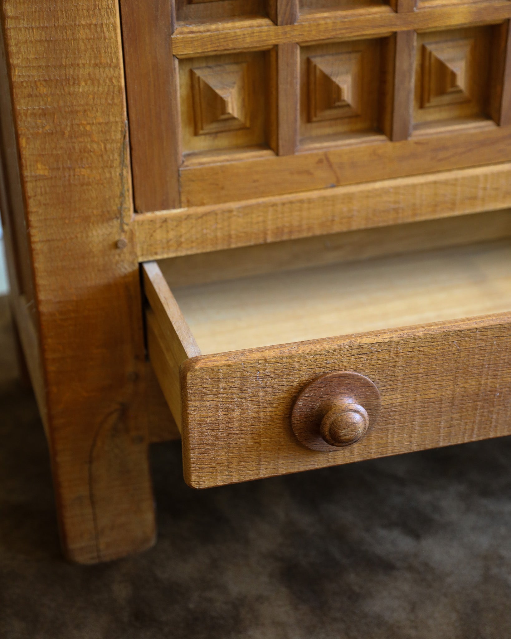 Close-up of a wooden furniture piece with a partially pulled-out drawer, showing a medium brown wood finish, carved panel details, and a round wooden knob.