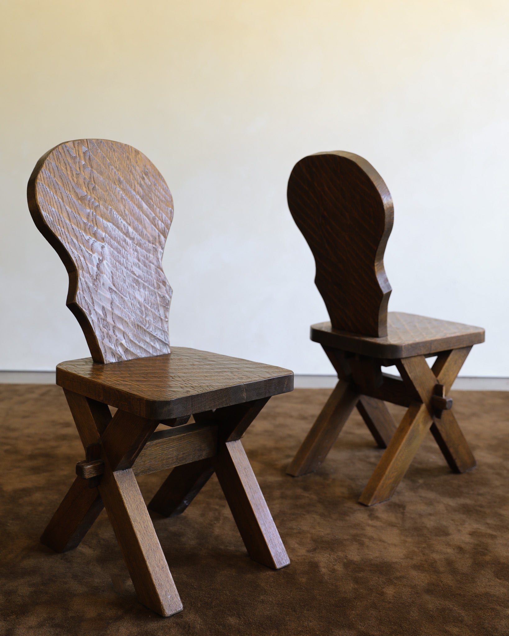 Two wooden chairs with uniquely shaped backs, shown on a brown carpet against a plain beige wall.