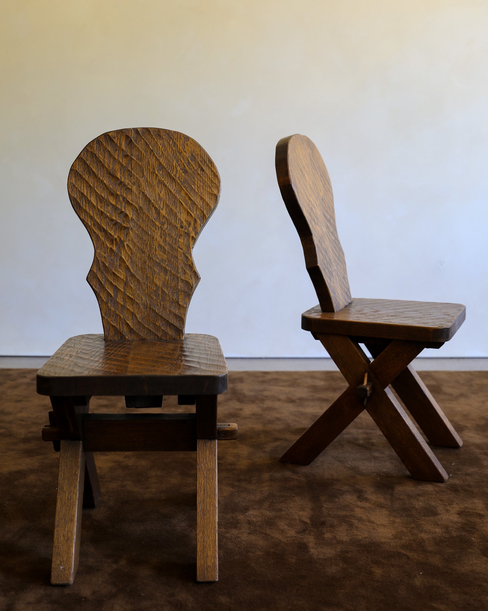Two rustic wooden chairs with carved backs, shown on a brown carpet against a plain off-white wall.