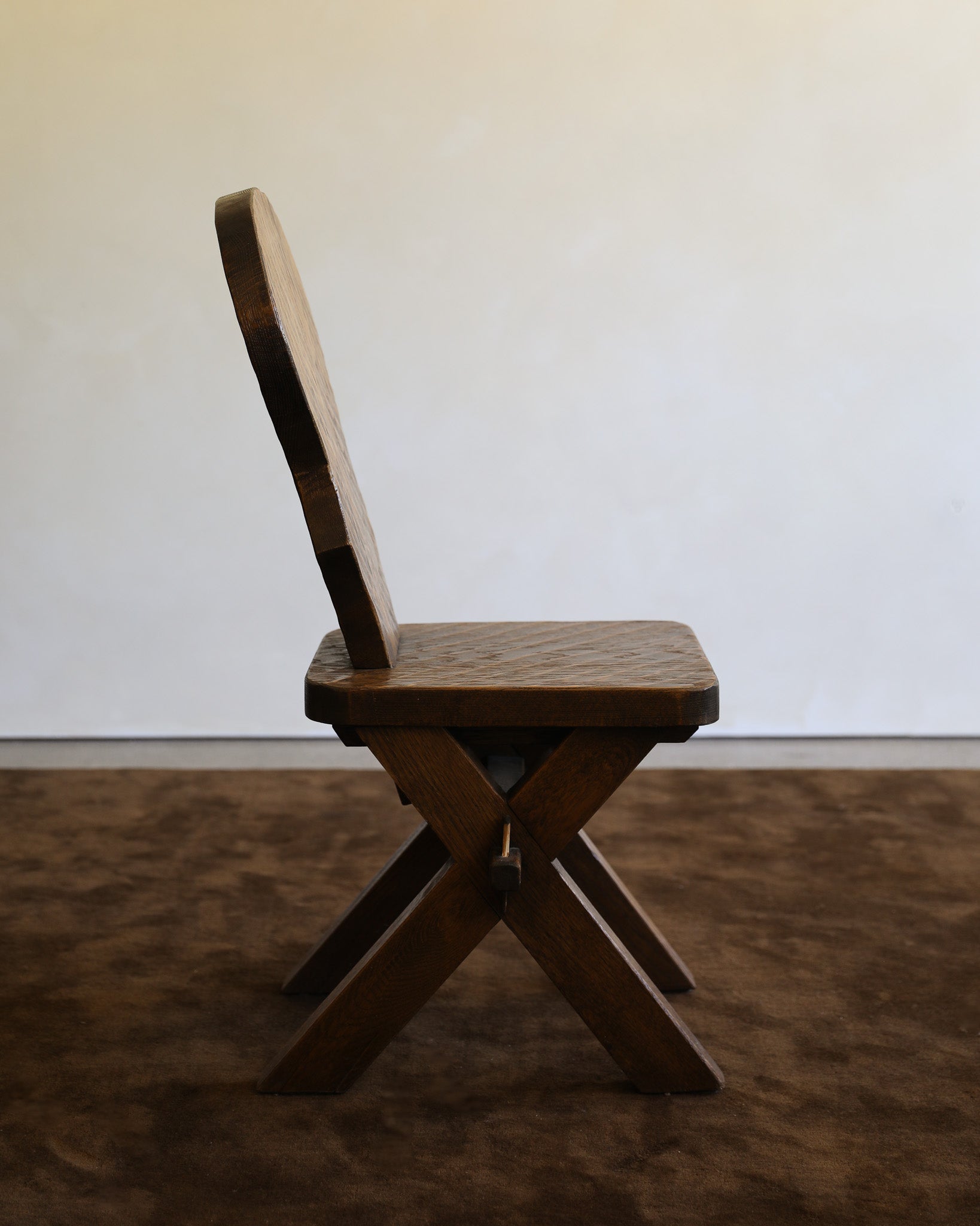A rustic wooden chair with a high curved backrest, shown from the side against a plain off-white wall and brown textured floor.